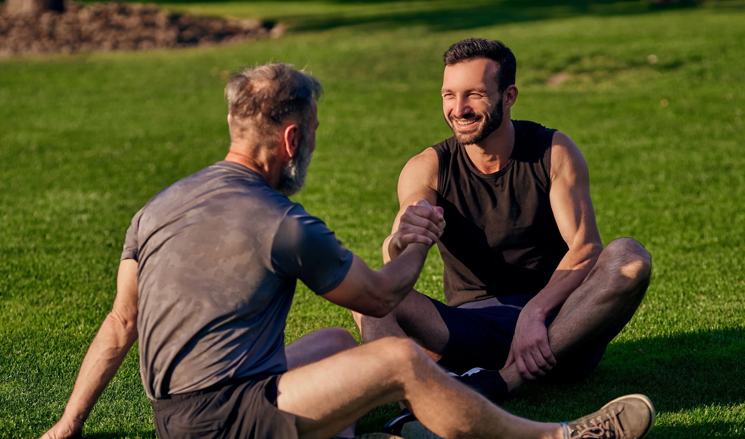 Two men sitting on grass, smiling, clasping hands in an arm wrestling or handshake gesture, outdoors on a sunny day.