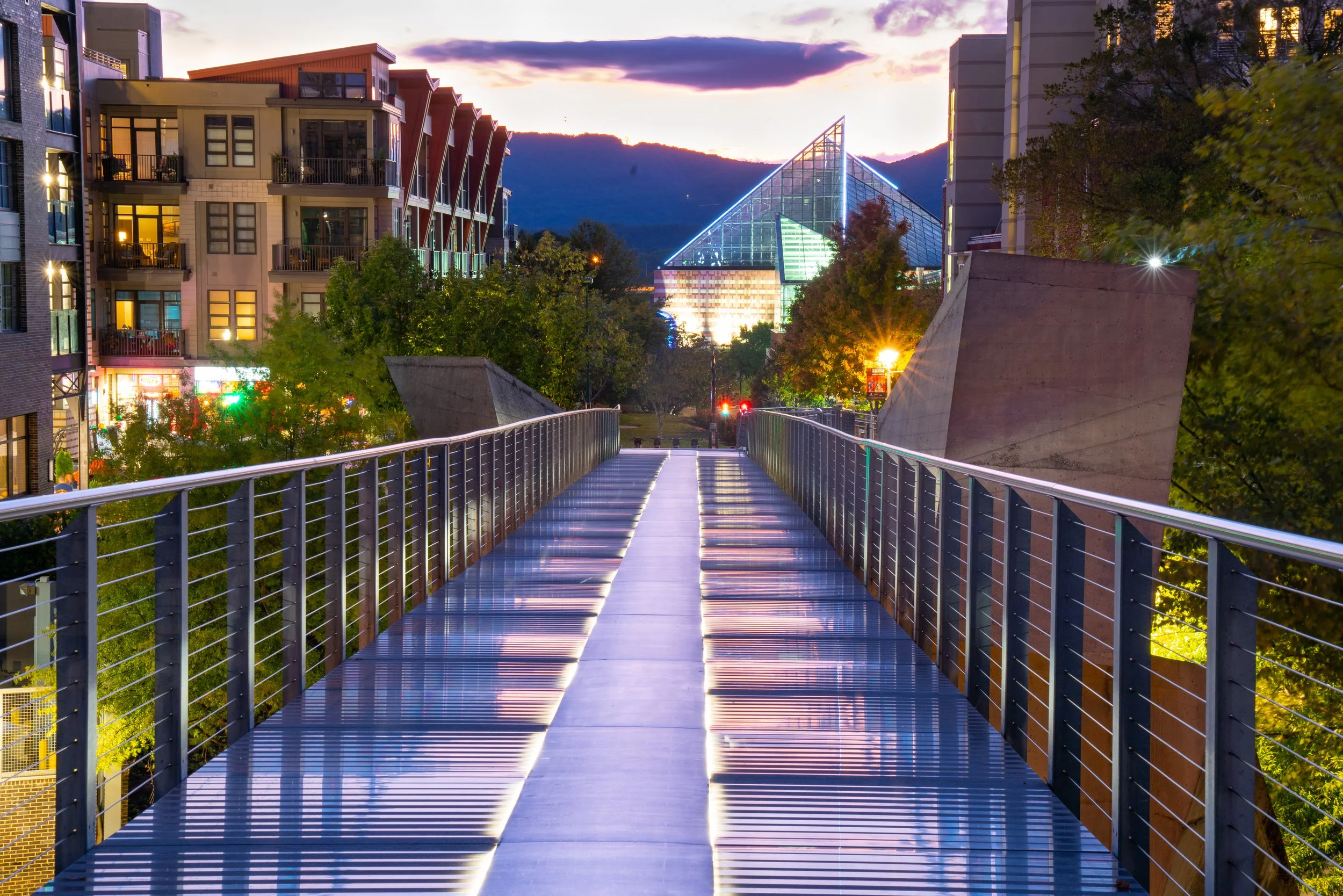 Aerial view of city buildings and a glass pyramid structure at sunset, with mountains in the background and a metal walking bridge in the foreground.