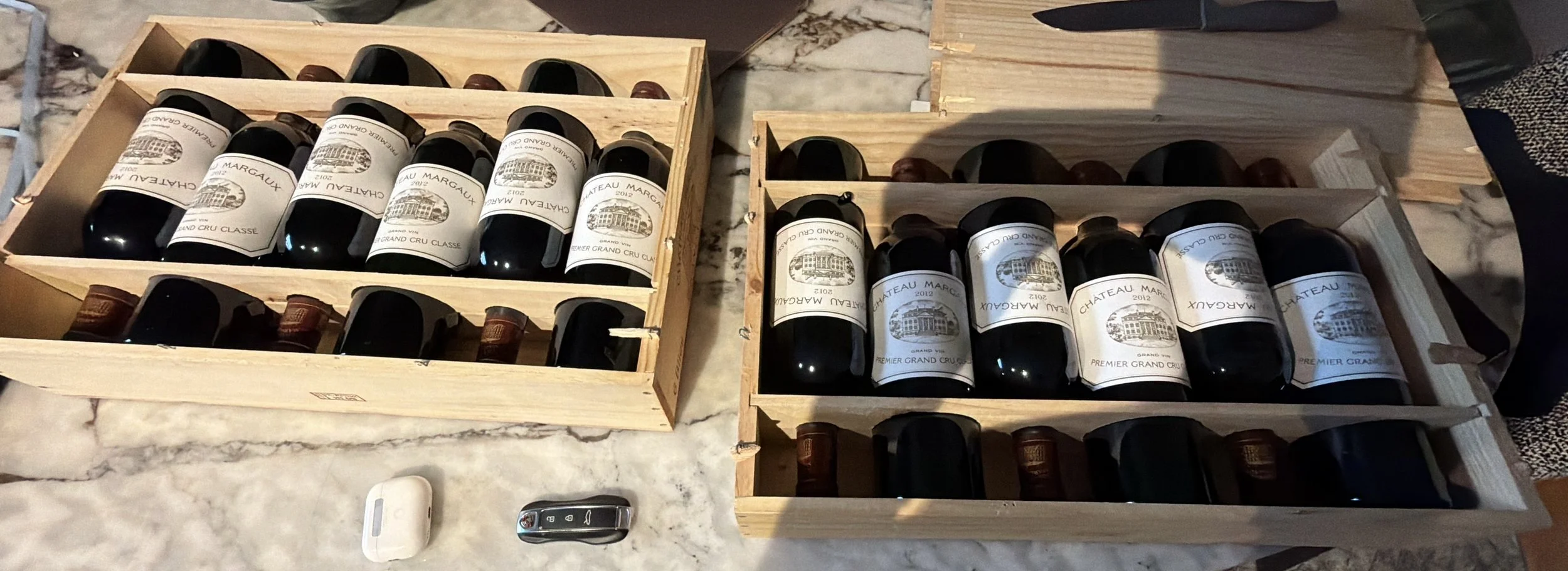 Deux caisses en bois contenant des bouteilles de vin rouge, avec des étiquettes indiquant Château Margaux 2012, posées sur un comptoir en marbre blanc, avec une clé de voiture et un objet blanc à côté.