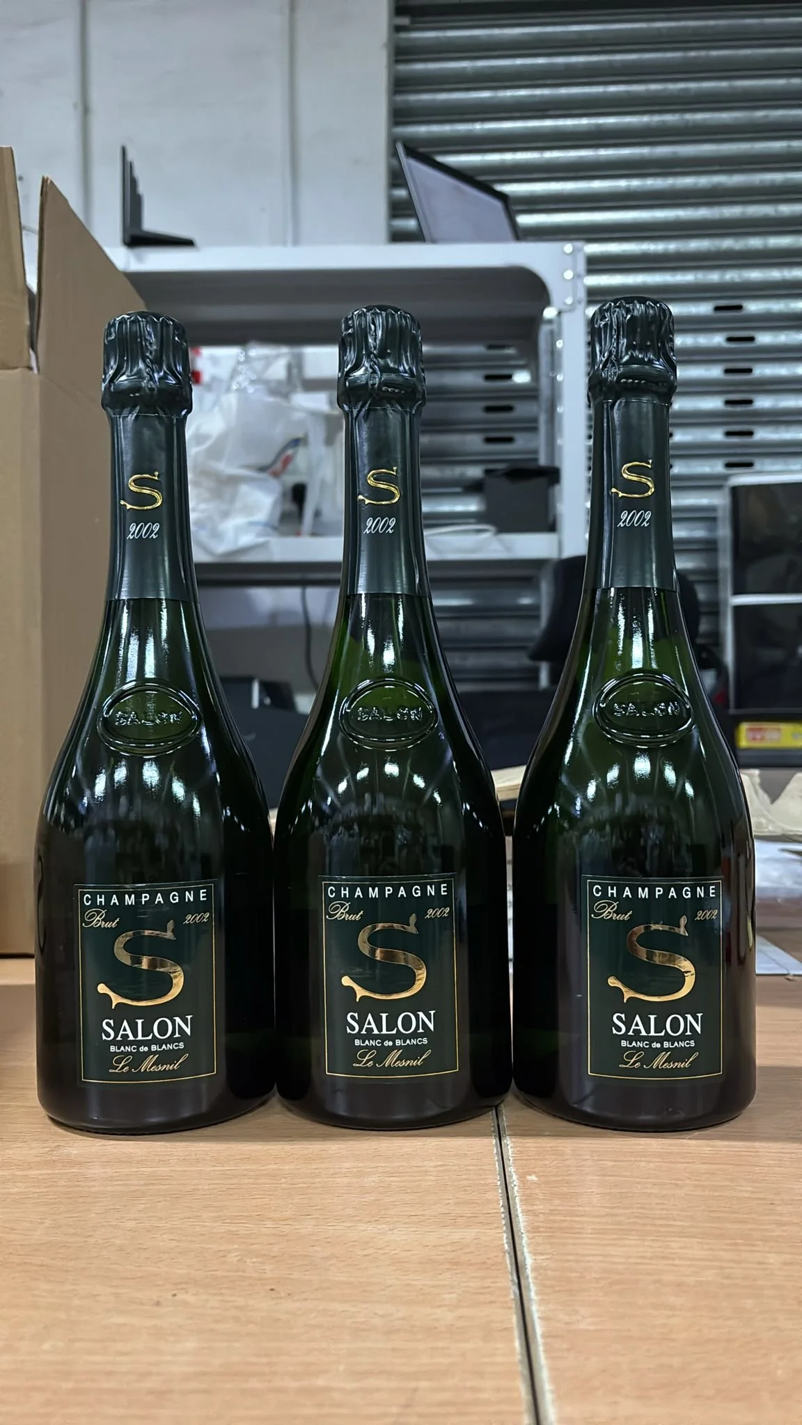 Trois bouteilles de champagne brutes, année 2002, positionnées sur une table en bois, avec des cartons et un arrière-plan industriel.
