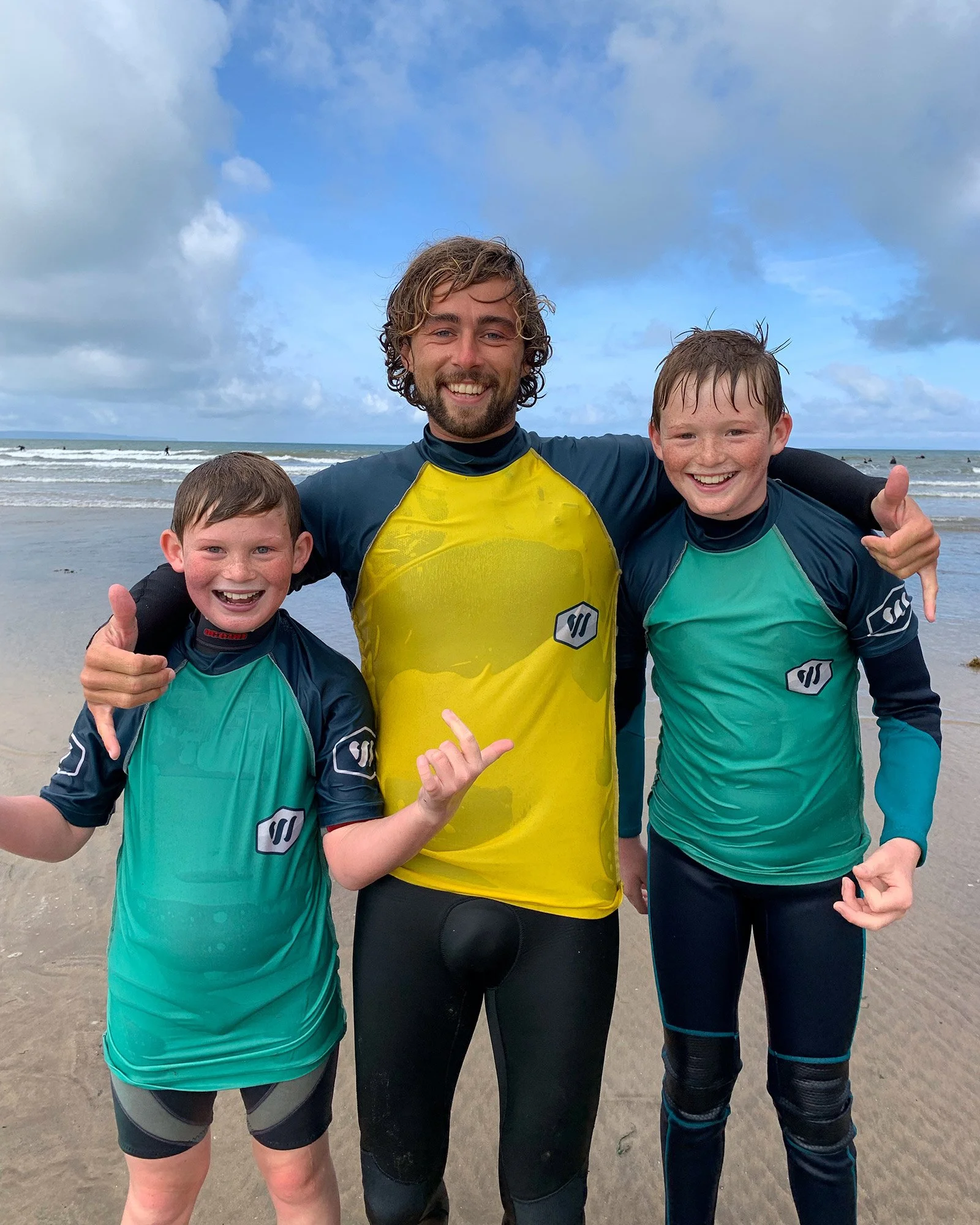 Three smiling individuals, two children and a man, in wetsuits at the beach, with ocean waves and a cloudy sky in the background.