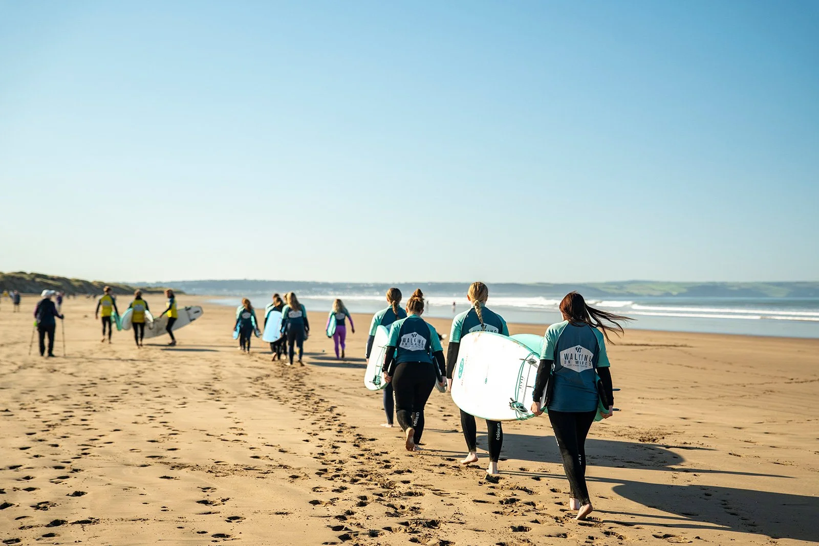 A group of people in wetsuits walking along a sandy beach while carrying surfboards, under a clear blue sky.