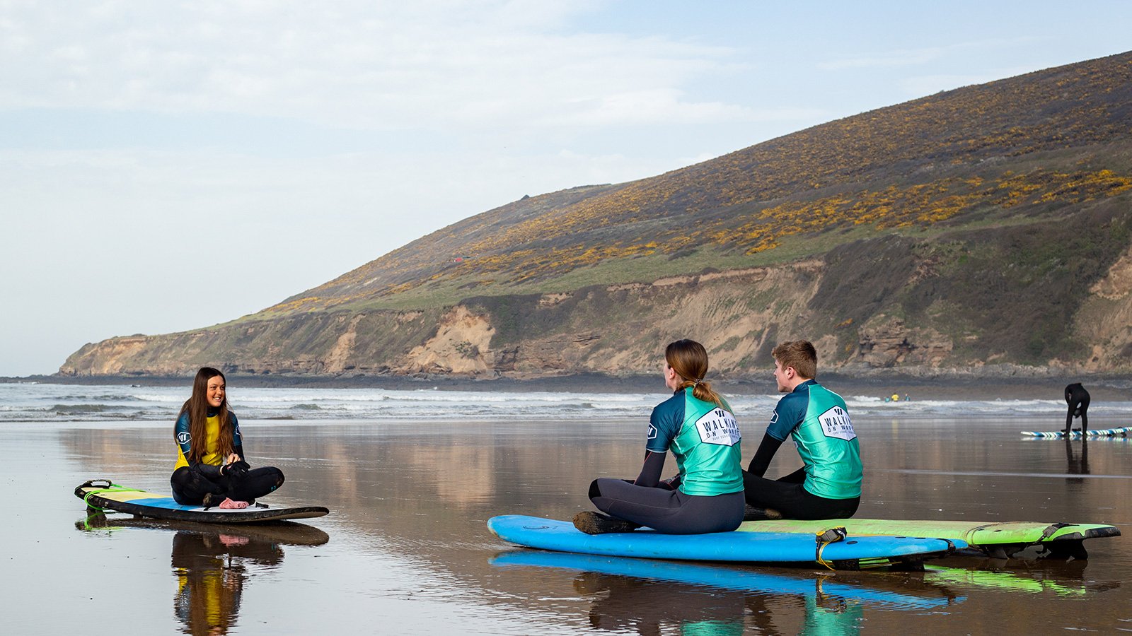 Three people sitting on surfboards at the beach, talking and smiling; two women and one man, with a scenic coastal landscape in the background.