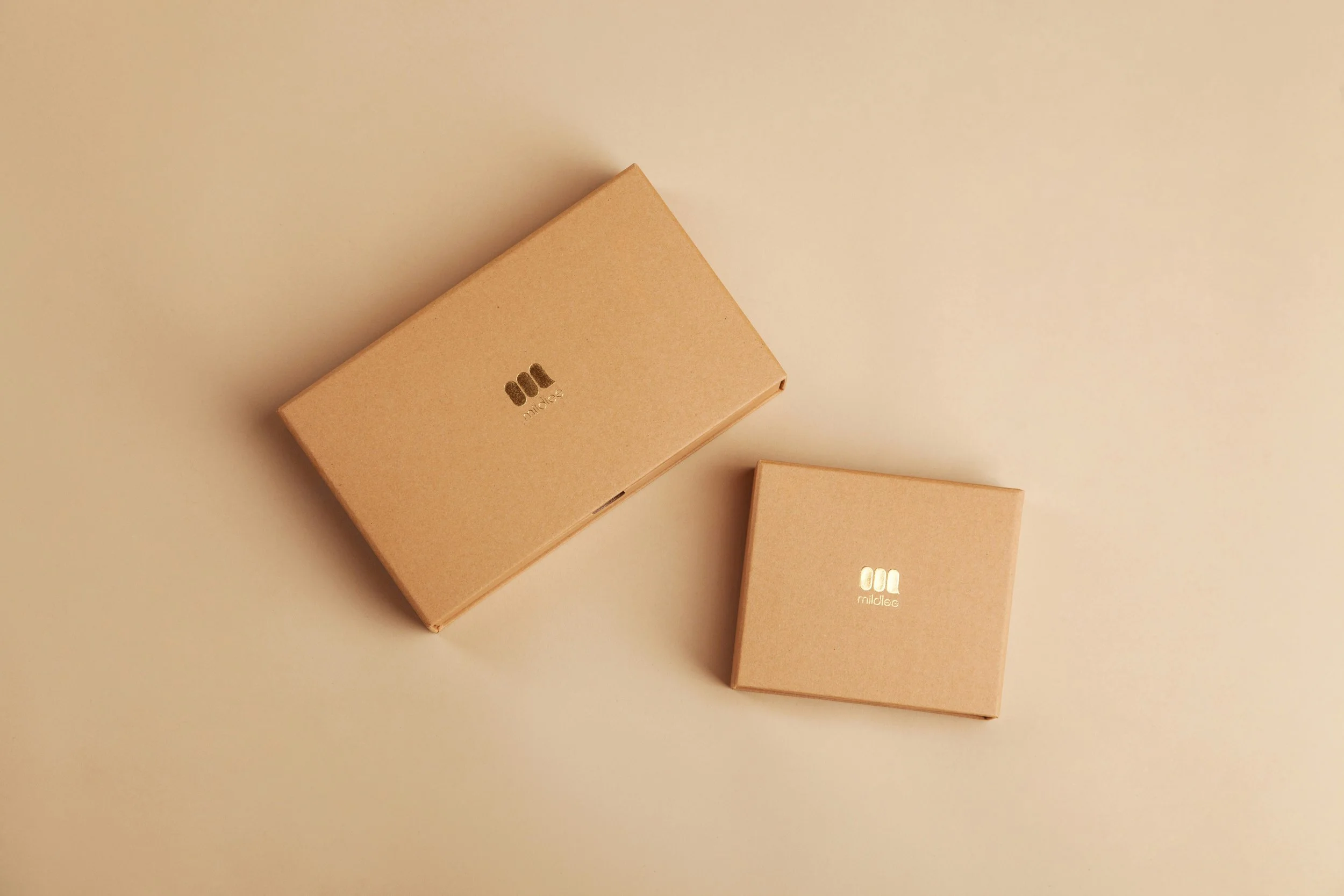 Two small beige boxes with a logo and the name 'mrailtee' printed in silver on the top, placed on a light beige background. One box is larger and positioned to the left, and a smaller box is to the right.