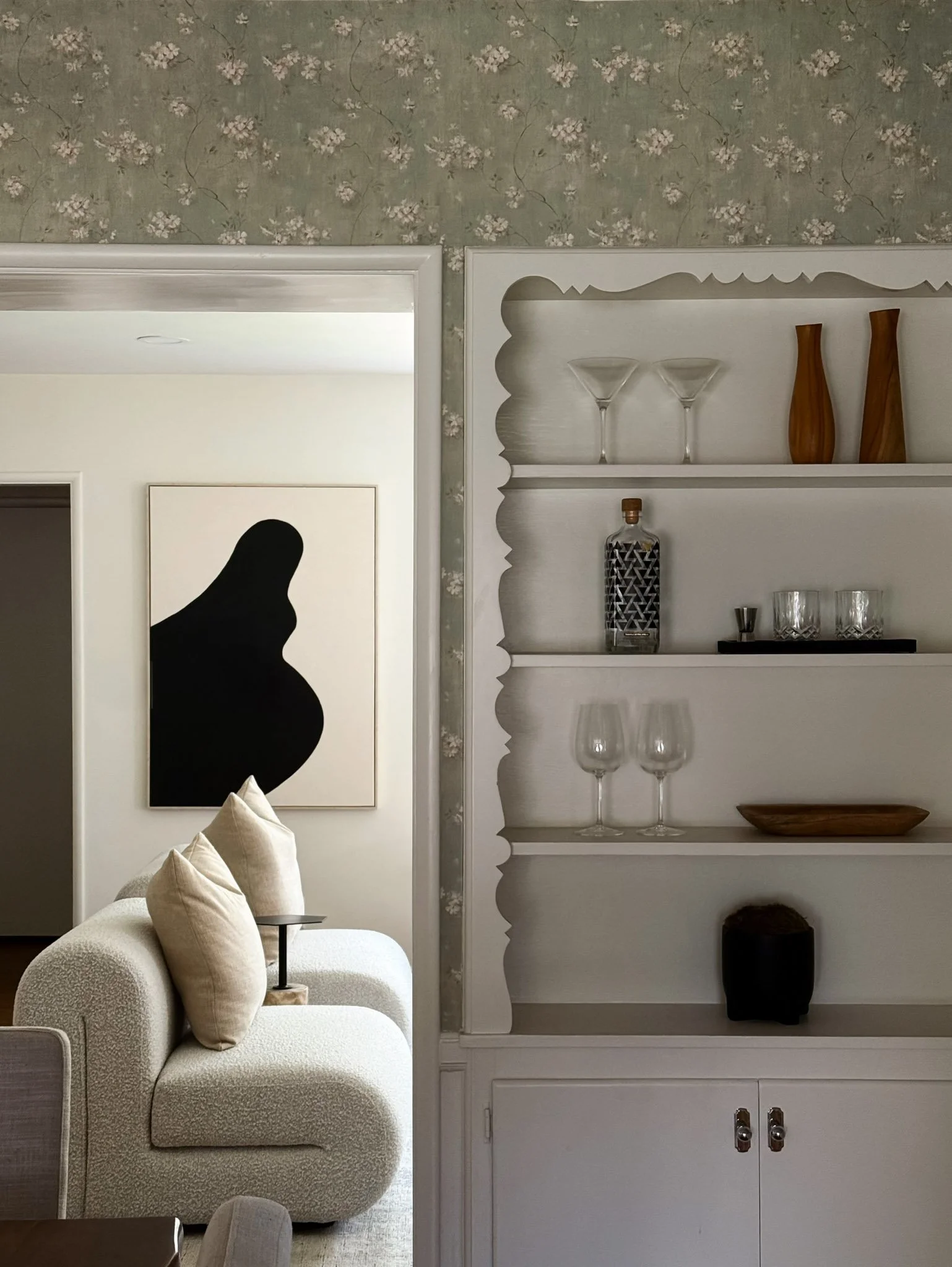 Living room with white textured sofa, beige pillows, abstract black and white portrait, white built-in shelf with glassware and wooden decorations, floral wallpaper, doorways, and modern decor.