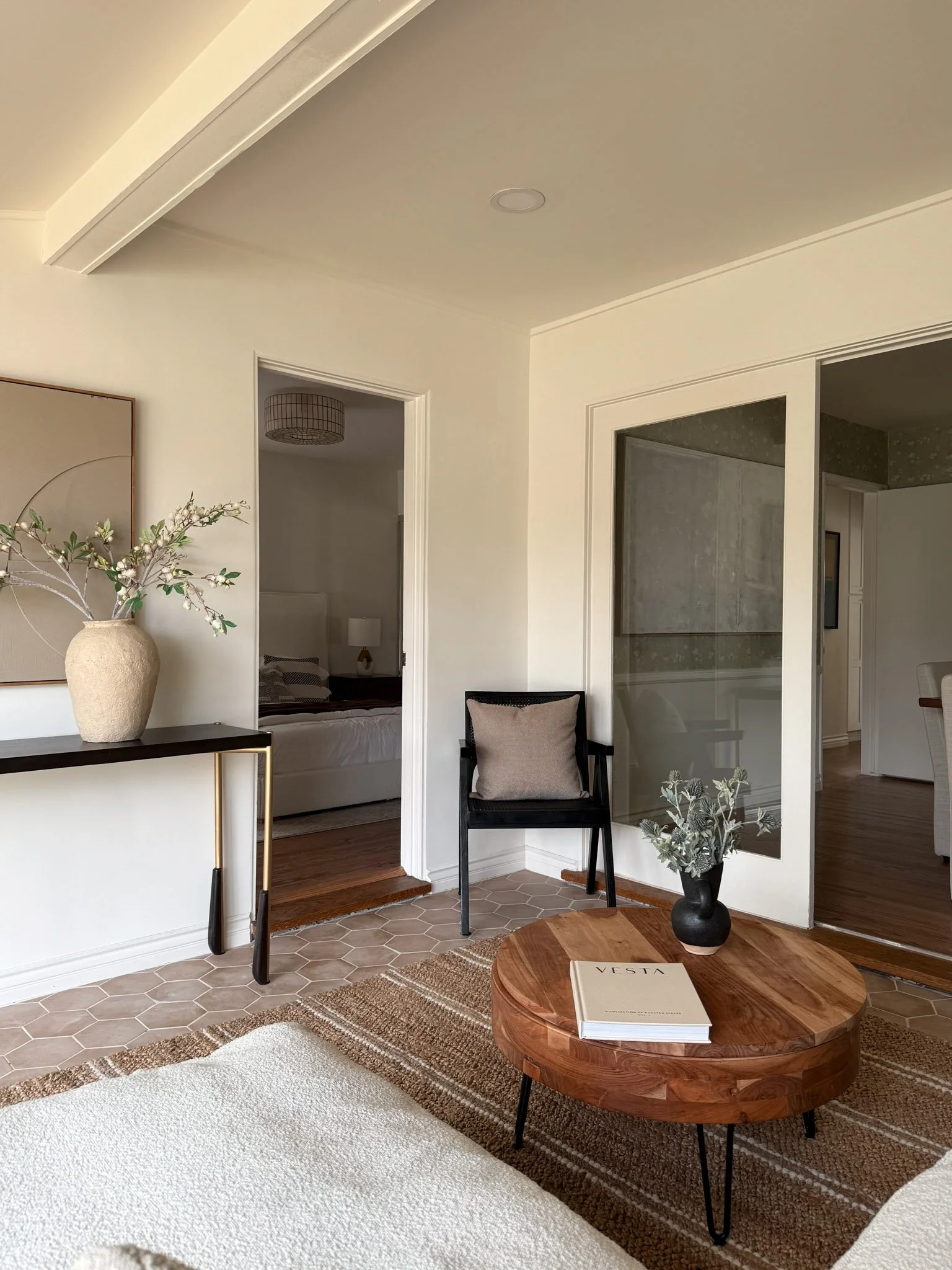 Interior of a modern living room with a round wooden coffee table, a beige and black armchair, a vase with greenery, and a textured wall with a glass divider. A bedroom is visible through an open doorway.