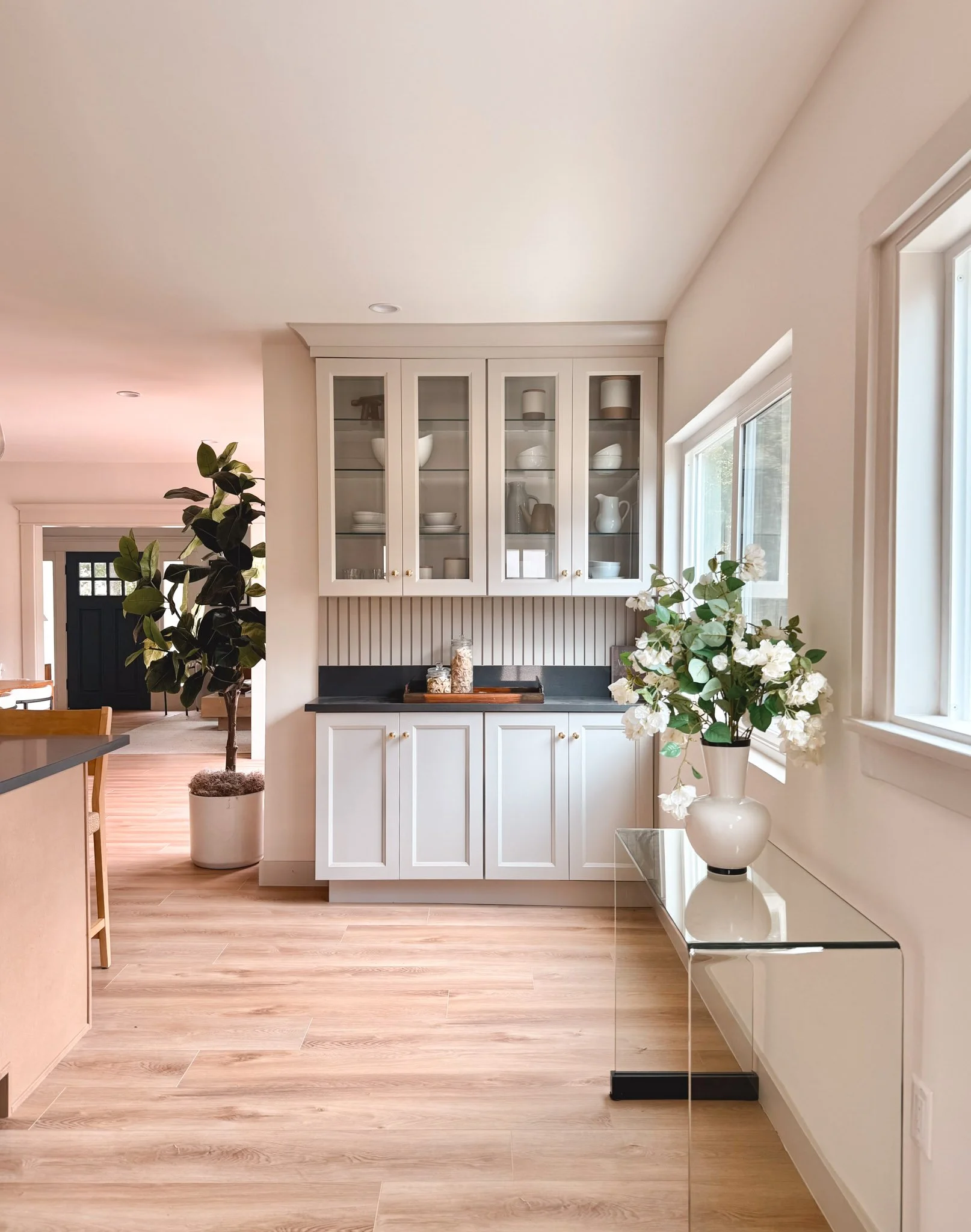 Bright kitchen area with white cabinets and glass doors, a black countertop, a large window with natural light, a white flower vase on a mirrored table, and a potted plant in the background.