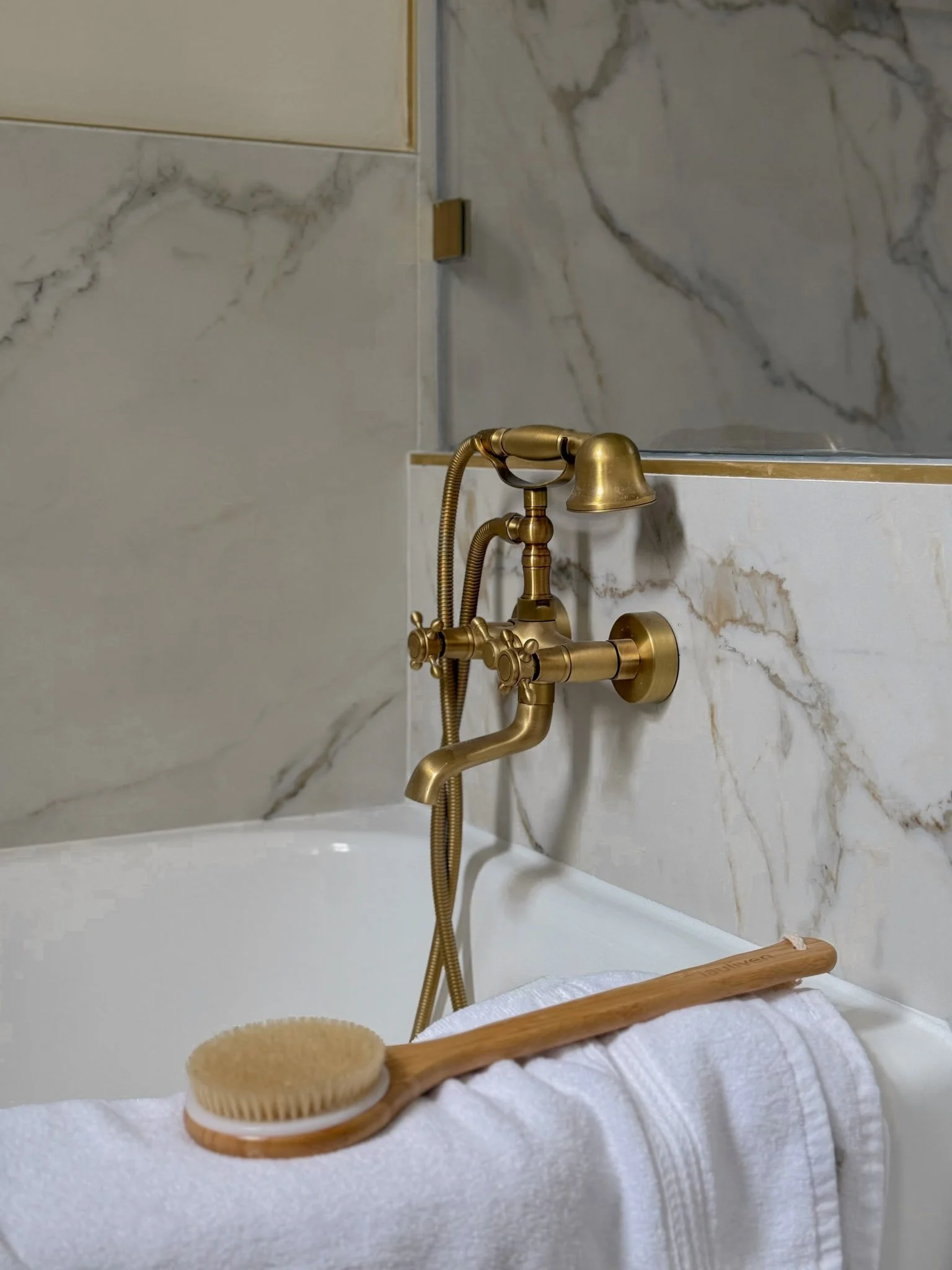 A white bathtub with a gold vintage-style faucet and handheld showerhead, a white towel, and a wooden bath brush resting on the towel, set against a marble wall.