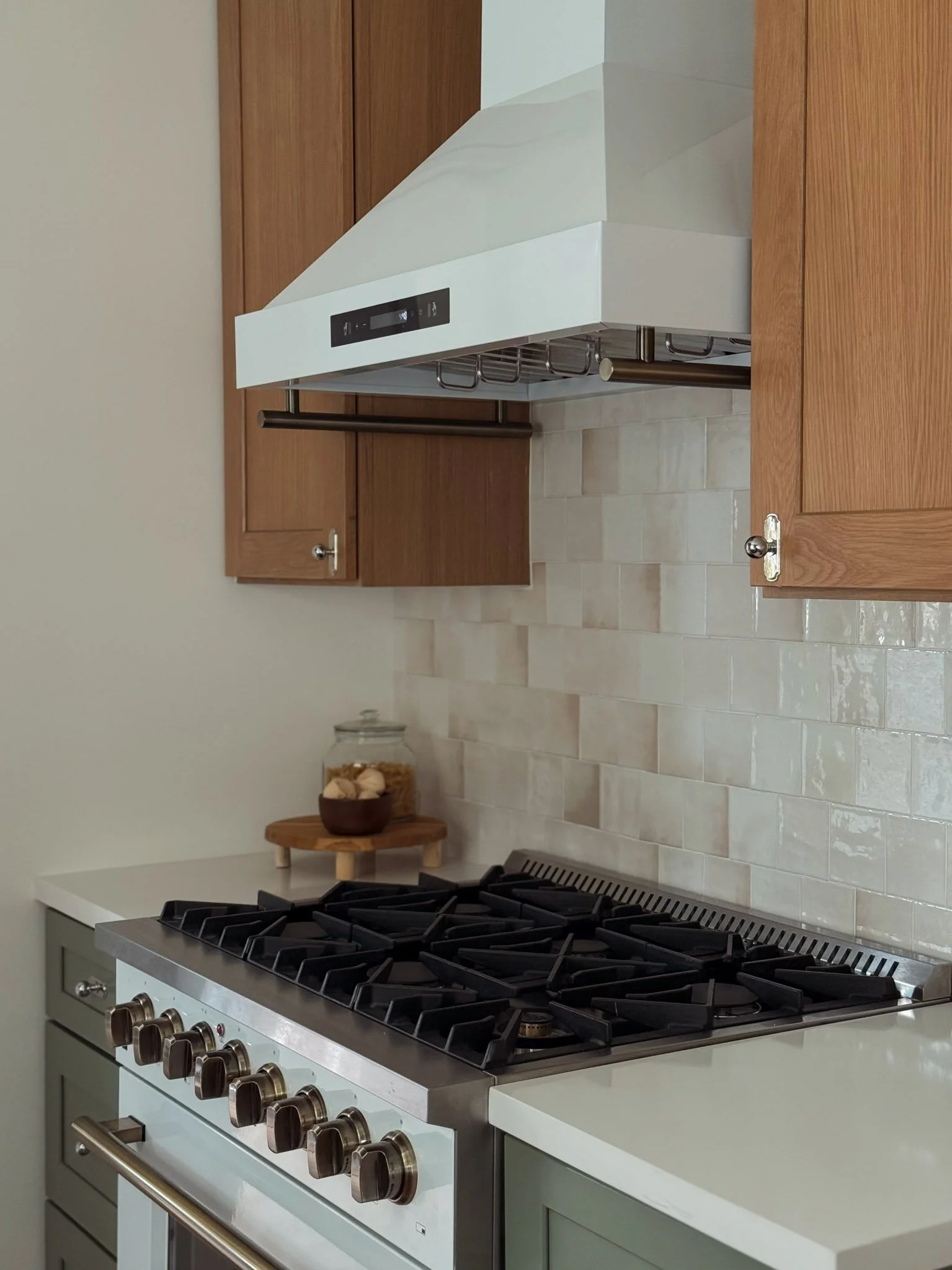 A modern kitchen stove with six burners, stainless steel finish, brown knobs, surrounded by white countertops, with wooden cabinets and a tiled beige backsplash in the background.