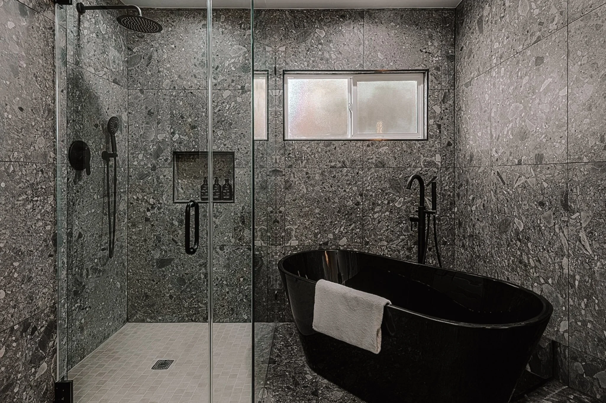 Modern bathroom with gray stone walls and floor, a black freestanding bathtub with a towel draped over the side, and a walk-in shower with glass doors and black fixtures. There is a small frosted window on the wall.