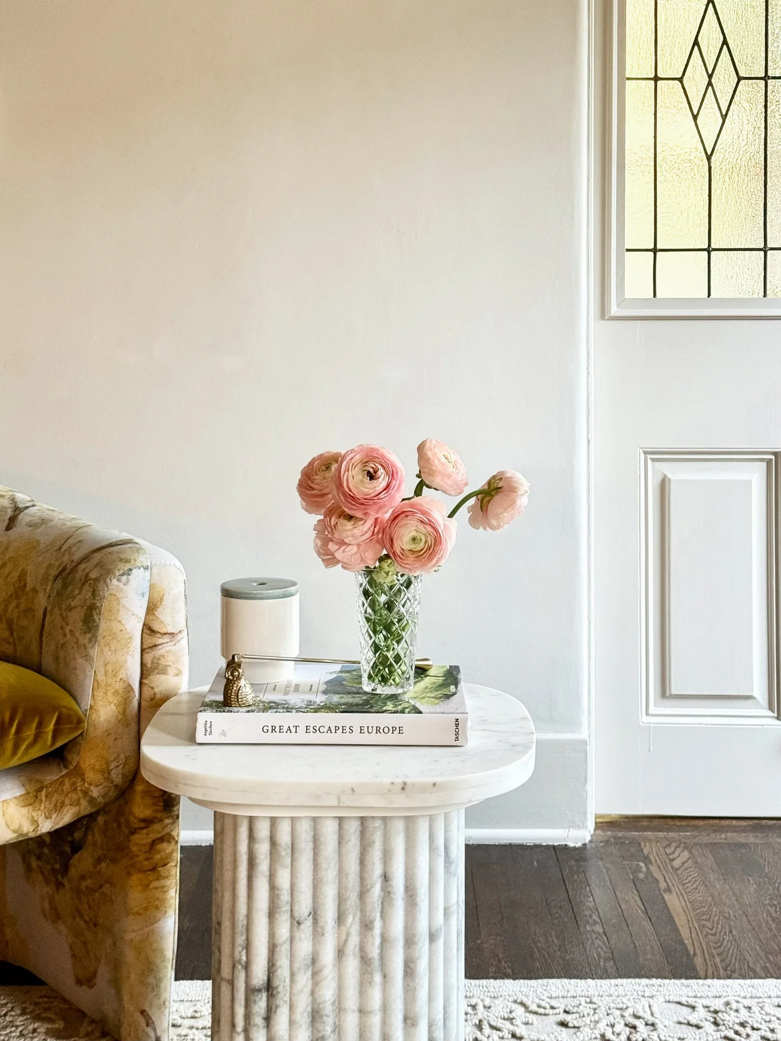 A round, marble side table with a fluted base, holding a glass vase with pink ranunculus flowers, a white book titled 'GREAT ESCAPES EUROPE', a small metallic owl figurine, and a white cylindrical container. The table is next to a floral-patterned co