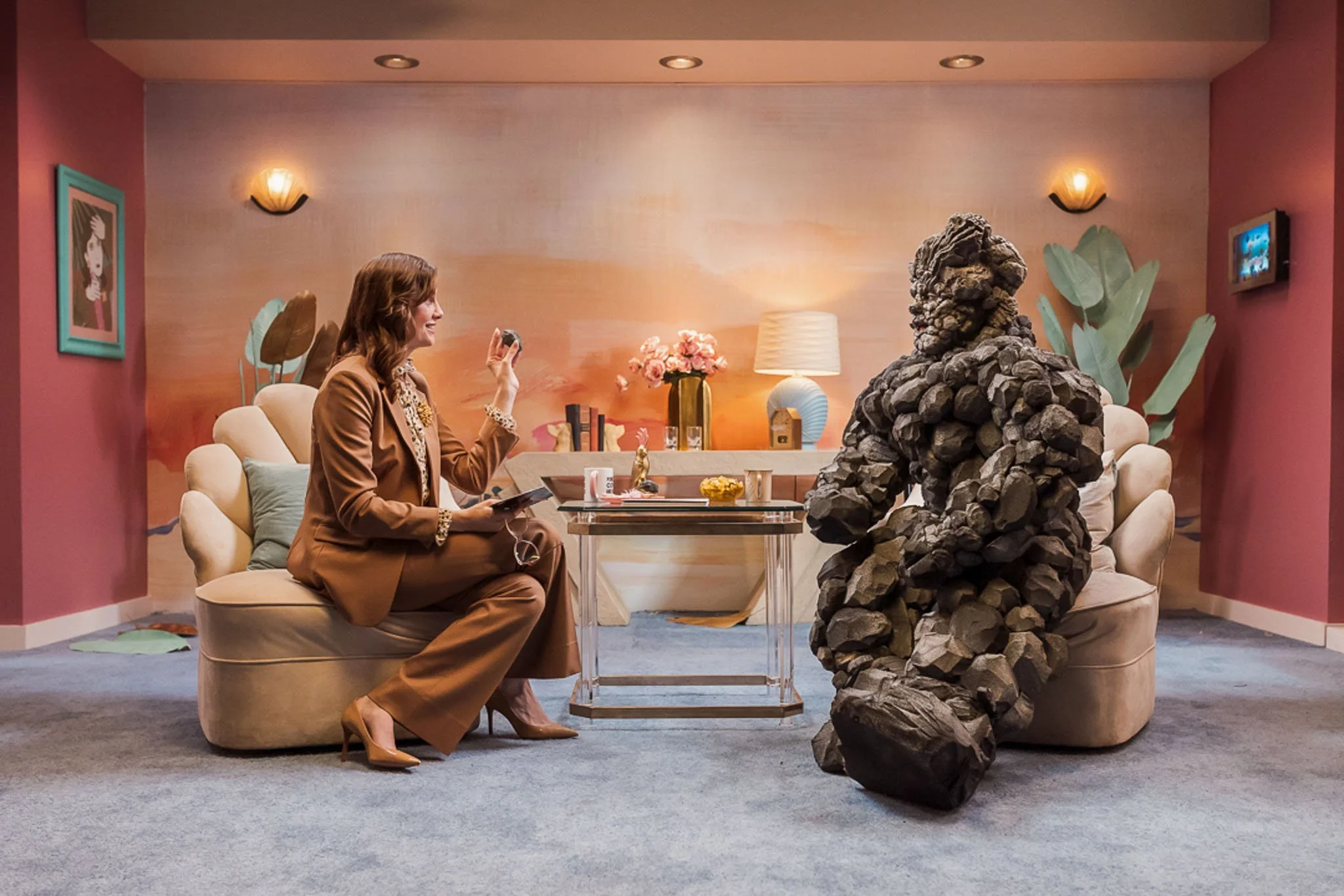 A woman in a brown suit sitting on a cream-colored sofa, talking to a large sculpture made of rocks in a decorated living room with pink and beige walls, a glass table, a lamp, flowers, and framed pictures.