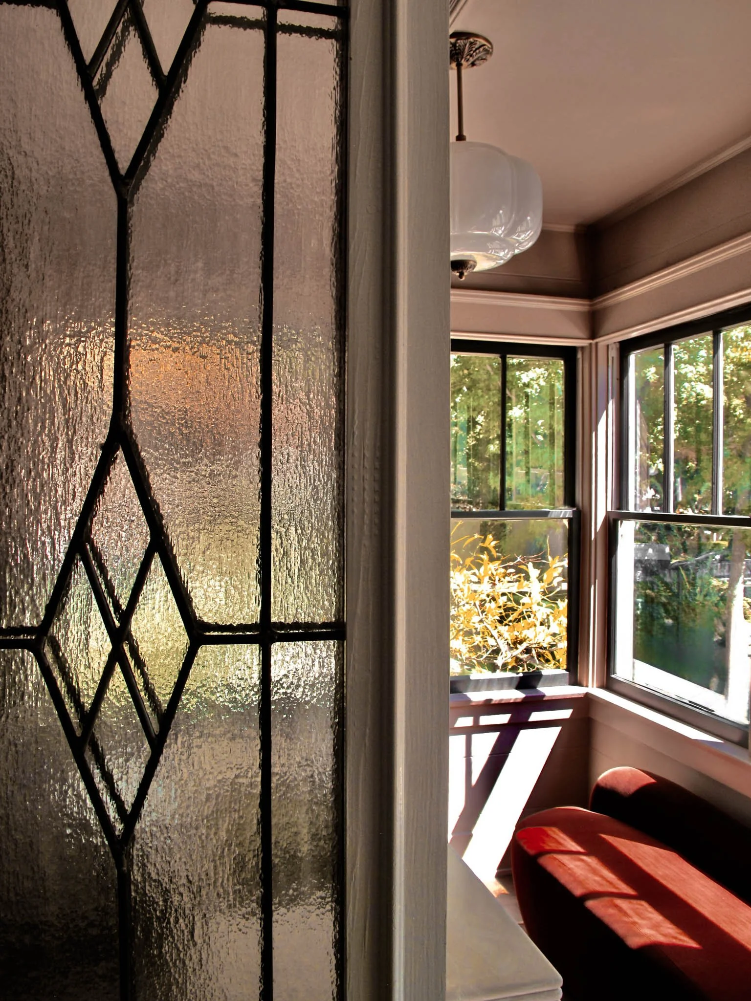 The image shows a glass door with textured glass and black geometric stained glass details on the left. Behind the door, part of a room is visible with large windows letting in natural sunlight, a hanging ceiling light, and a red cushioned bench or s