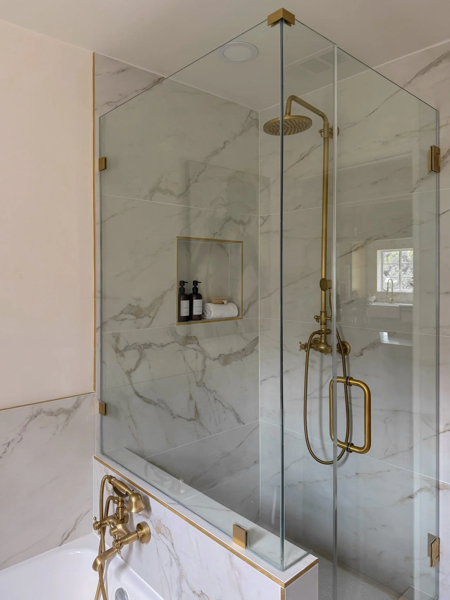 A modern walk-in shower with gold fixtures, glass enclosure, marble walls, and a built-in niche with shower products.
