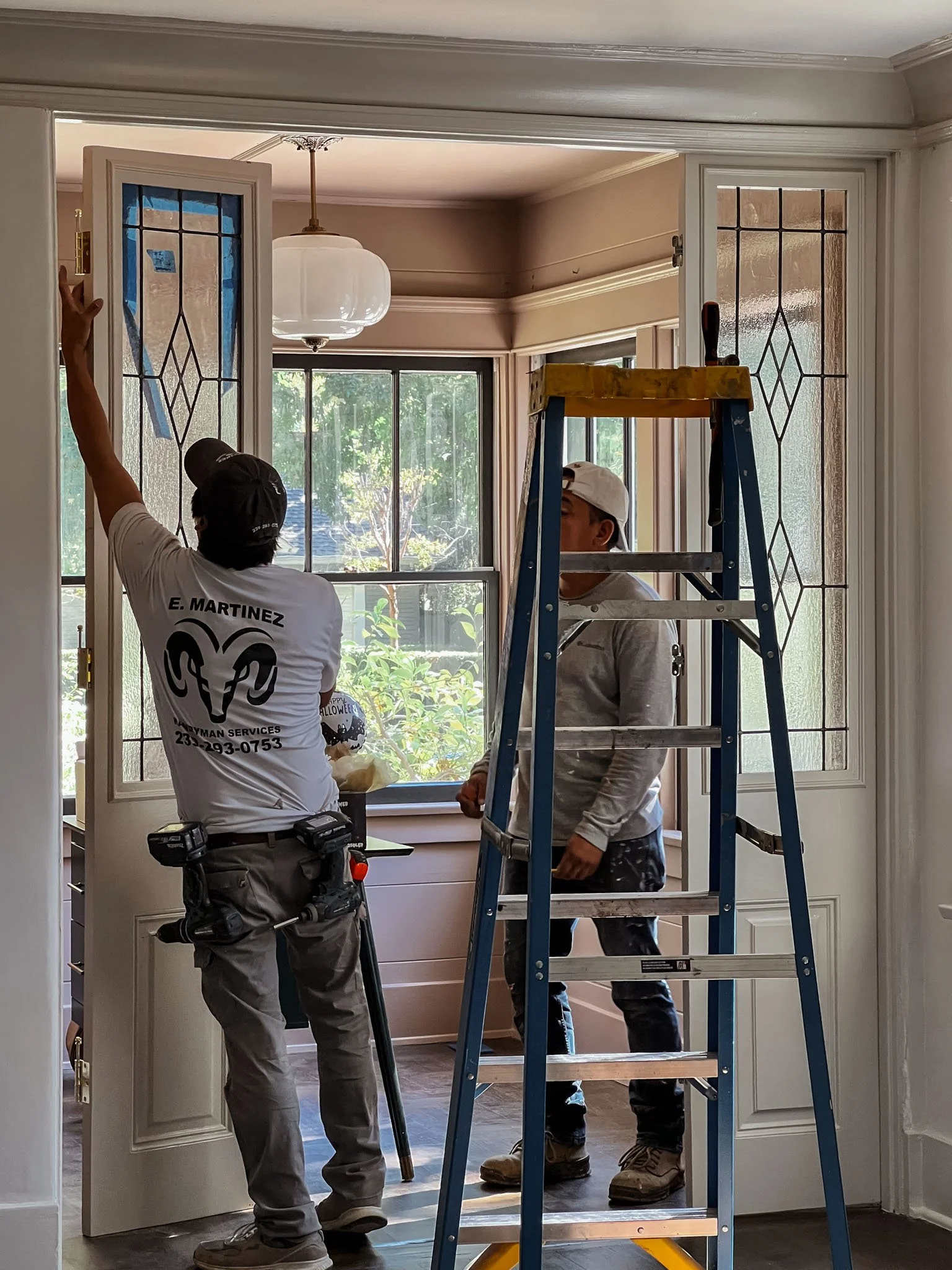 Two workers installing or repairing stained glass window panels in a door, with one on a ladder and the other standing nearby, inside a well-lit room.