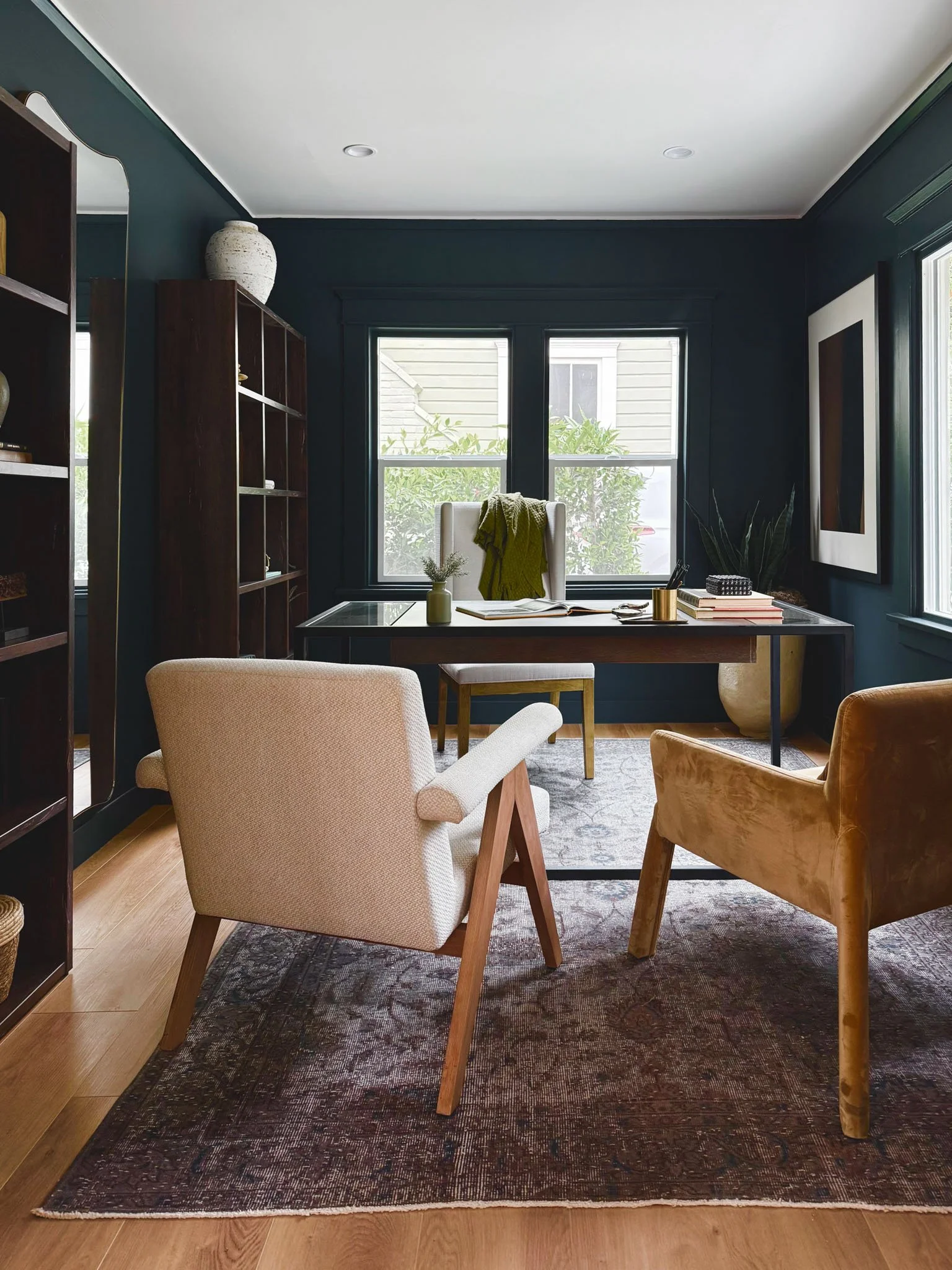 Home office with a dark green painted wall, a large window with outdoor view, a black desk with decorations, two chairs in front, and bookshelves.