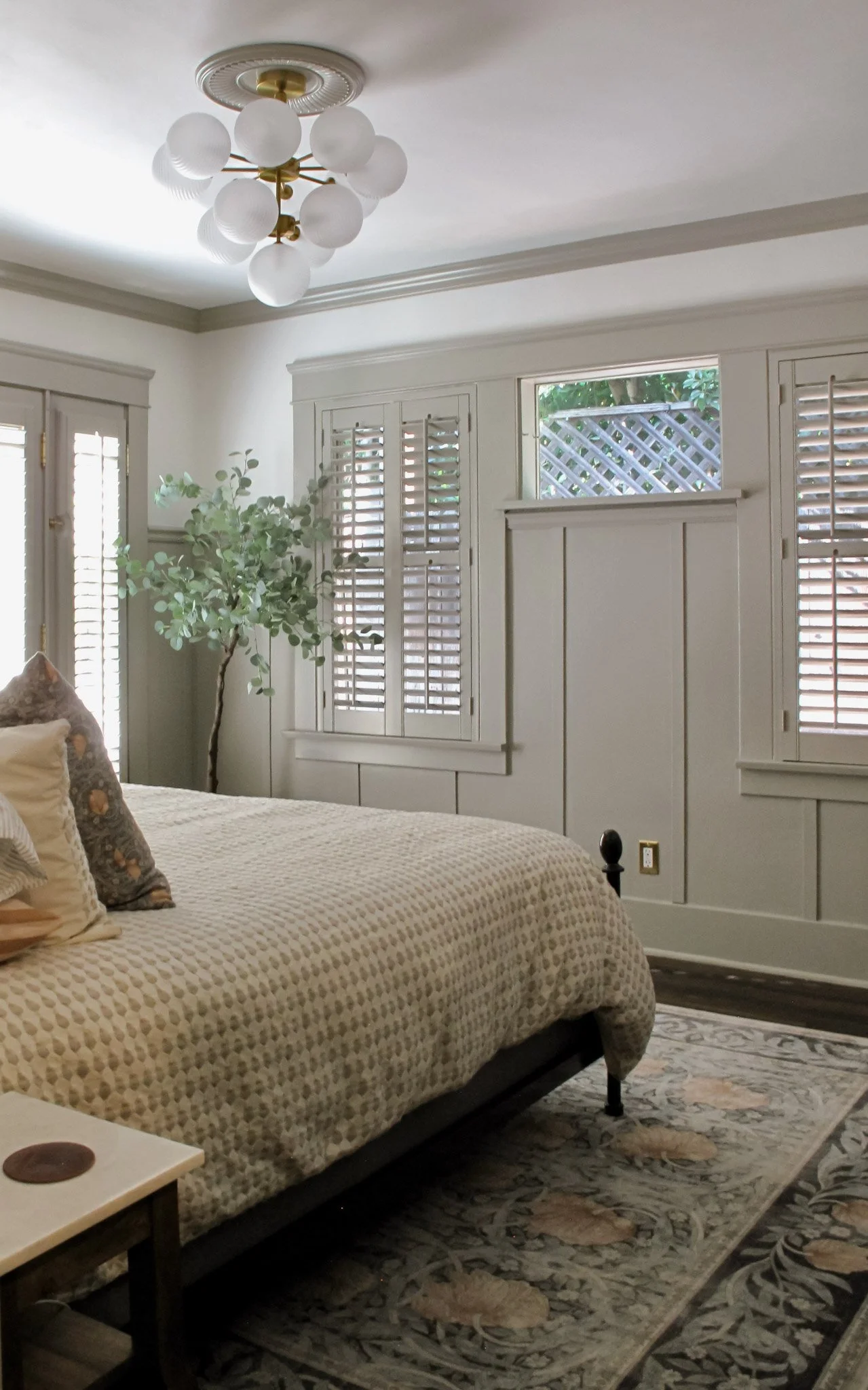 A bedroom with white walls, large windows with shutters, a bed with a beige patterned comforter, decorative pillows, and a rug on a wooden floor, a potted plant near the window, and a modern chandelier hanging from the ceiling.