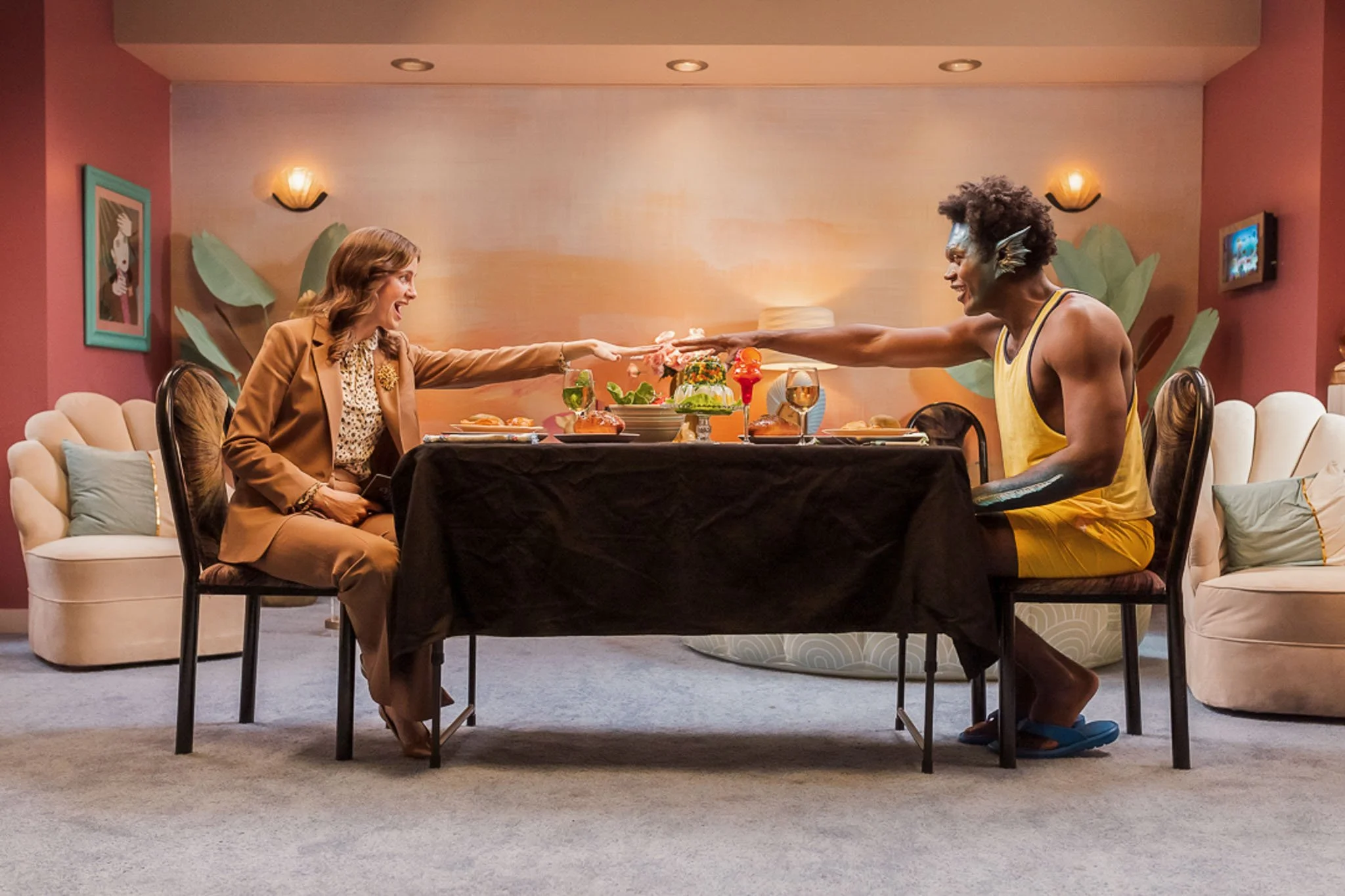 A woman and a man are reaching across a dining table towards each other, smiling, in a colorful, decorated room.