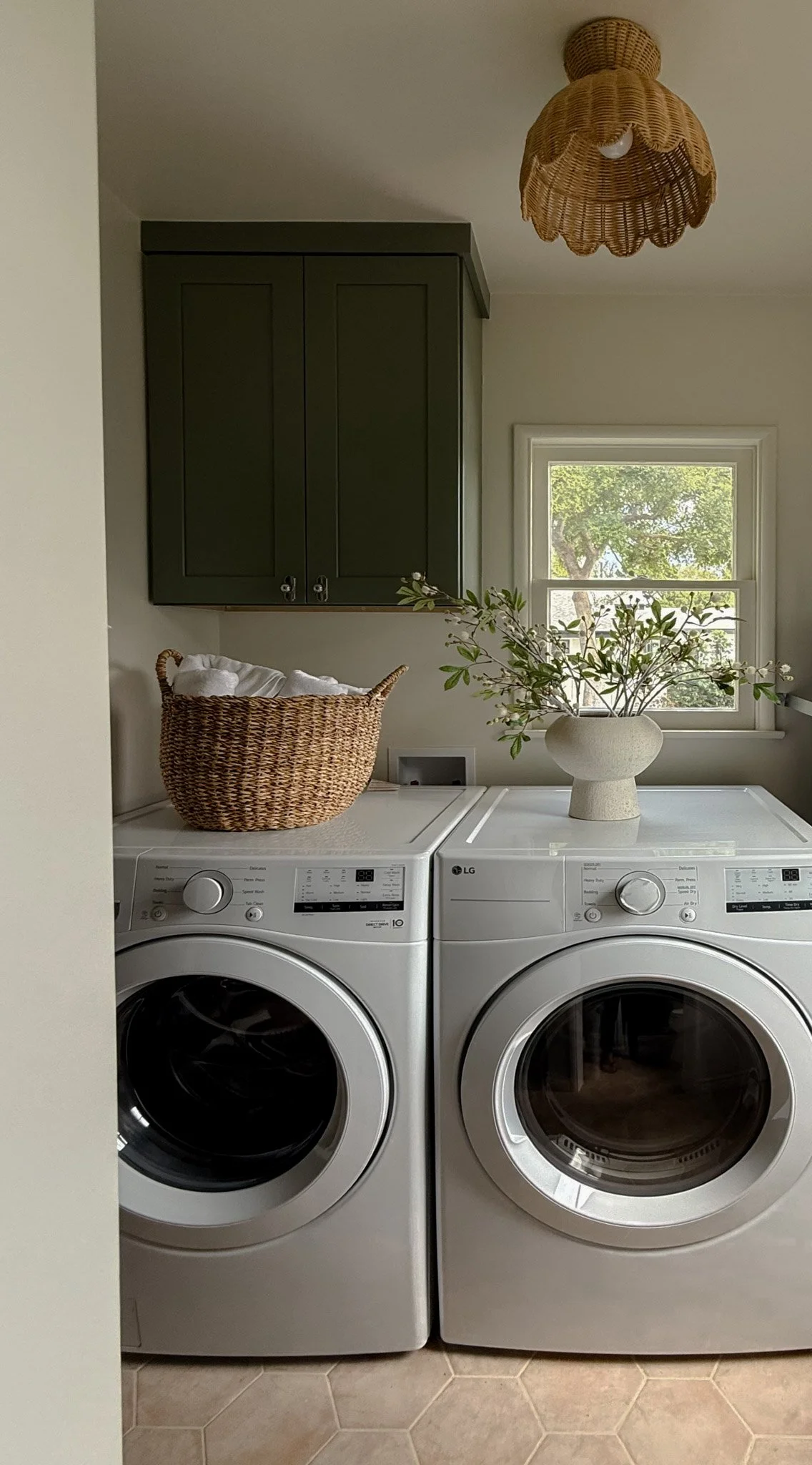 A laundry room with a front-loading washer and dryer, a wicker basket with folded towels on top, a vase with greenery, and a window with trees outside.