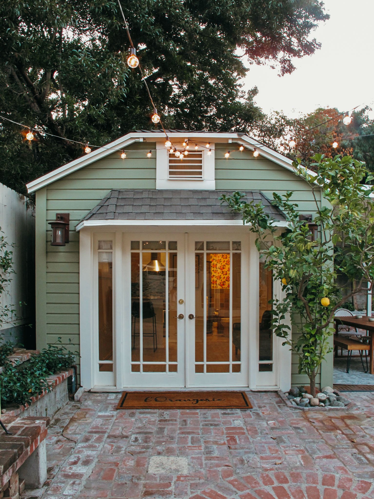 View of a small house with French doors, a brick patio in front, a lemon tree on the right, and string lights hanging above. The house has light green siding and a gable roof.