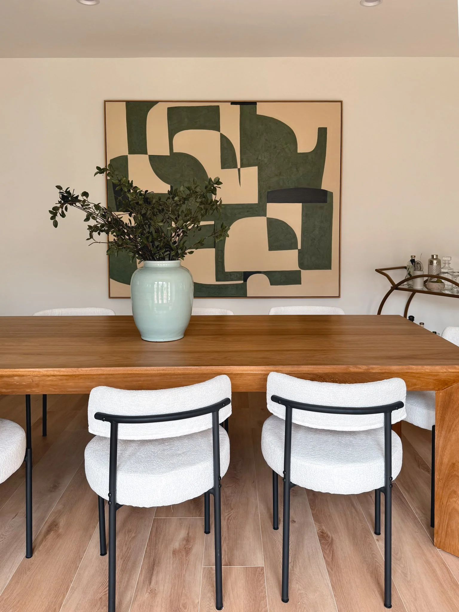 A dining room with a wooden table, six white upholstered chairs with black metal legs, a large abstract green and beige painting on the wall, a vase with greenery on the table, and a bar cart with bottles and barware to the side.