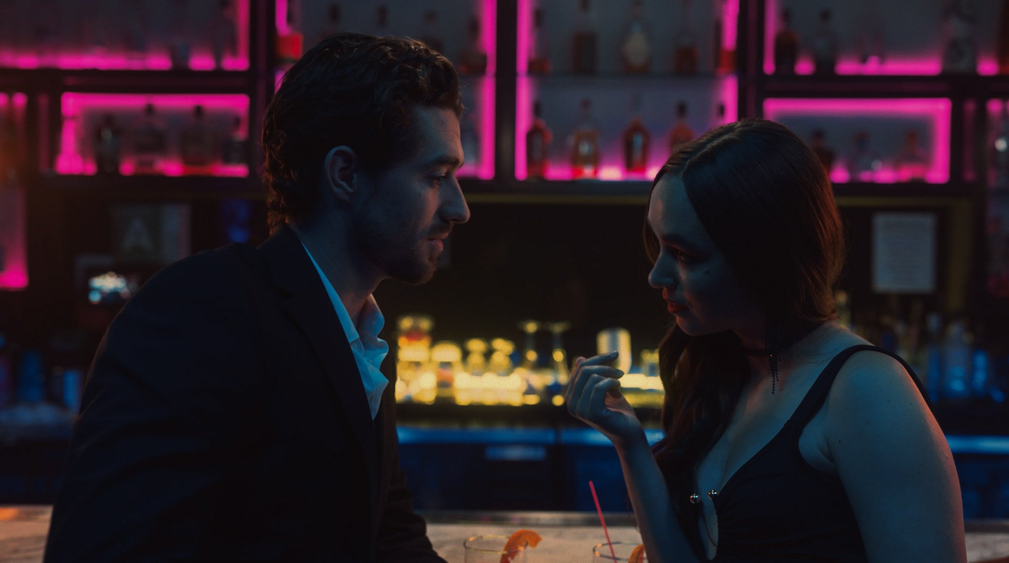 A man and woman sitting at a bar, close to each other, in a dimly lit setting with pink and purple neon lights in the background. The woman appears to be touching her face and the man is looking at her.