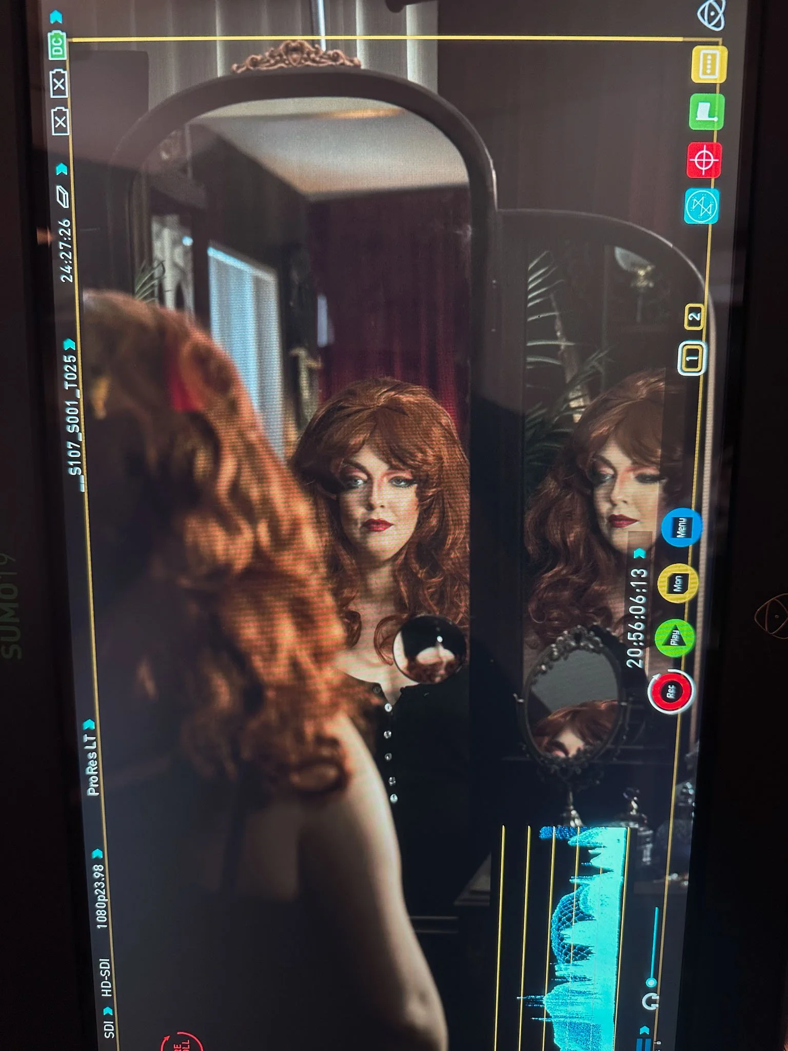 A woman with curly red hair and makeup looks into a mirror, with her back turned to the camera, in a dimly lit room with dark red curtains and plants in the background.