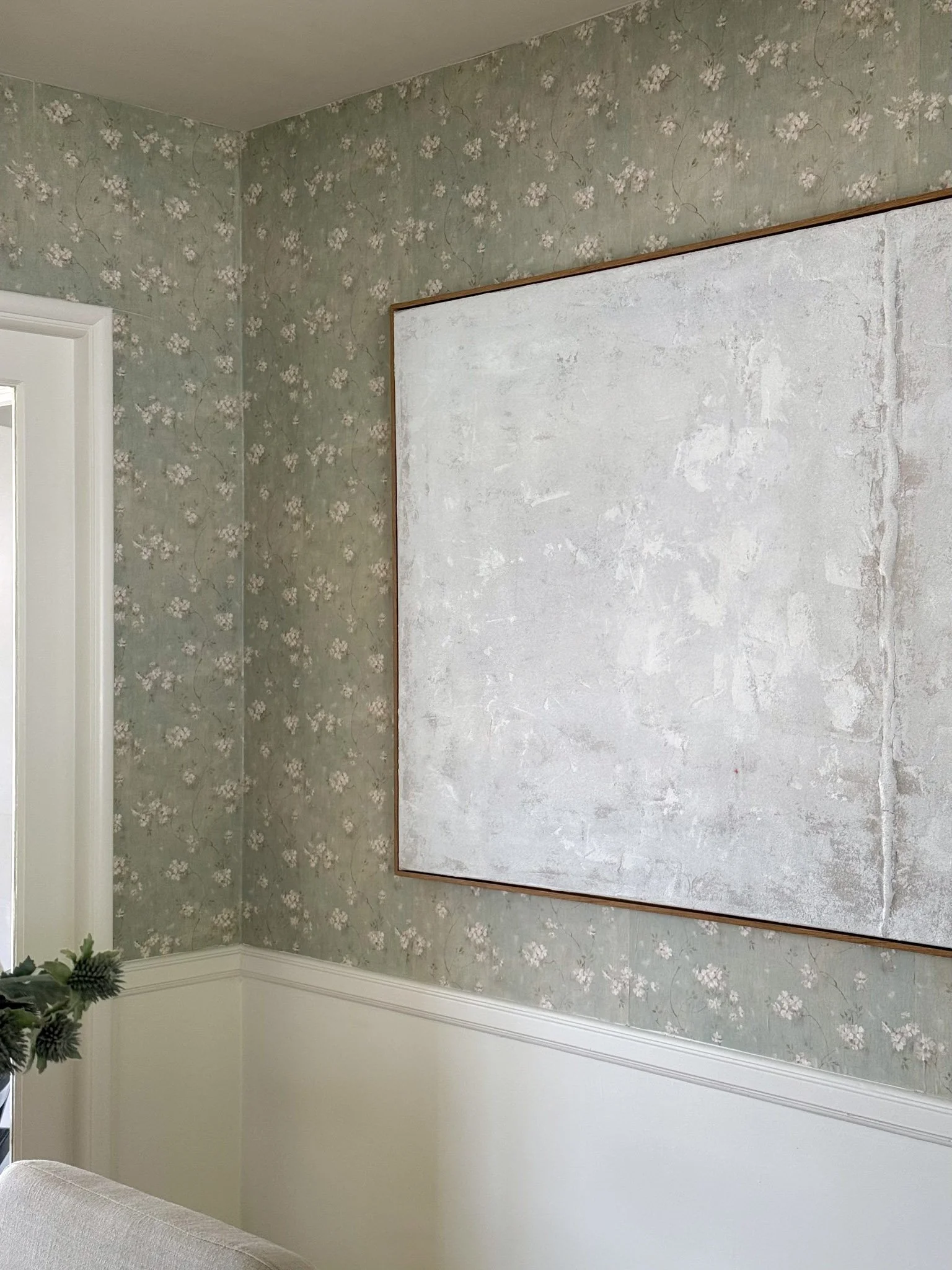 Empty wall with floral wallpaper partially covered by a white textured board, with a corner of a beige couch visible in the lower left.