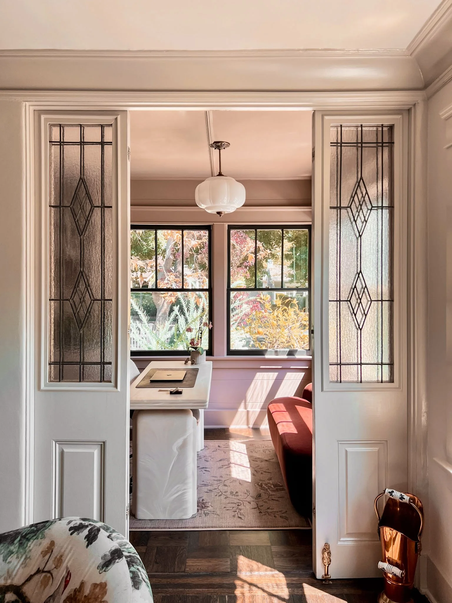 View through a classic white wooden and glass double door into a cozy sunlit room with a white desk, pink armchair, large windows showing trees and plants outside, and a hanging ceiling lamp.