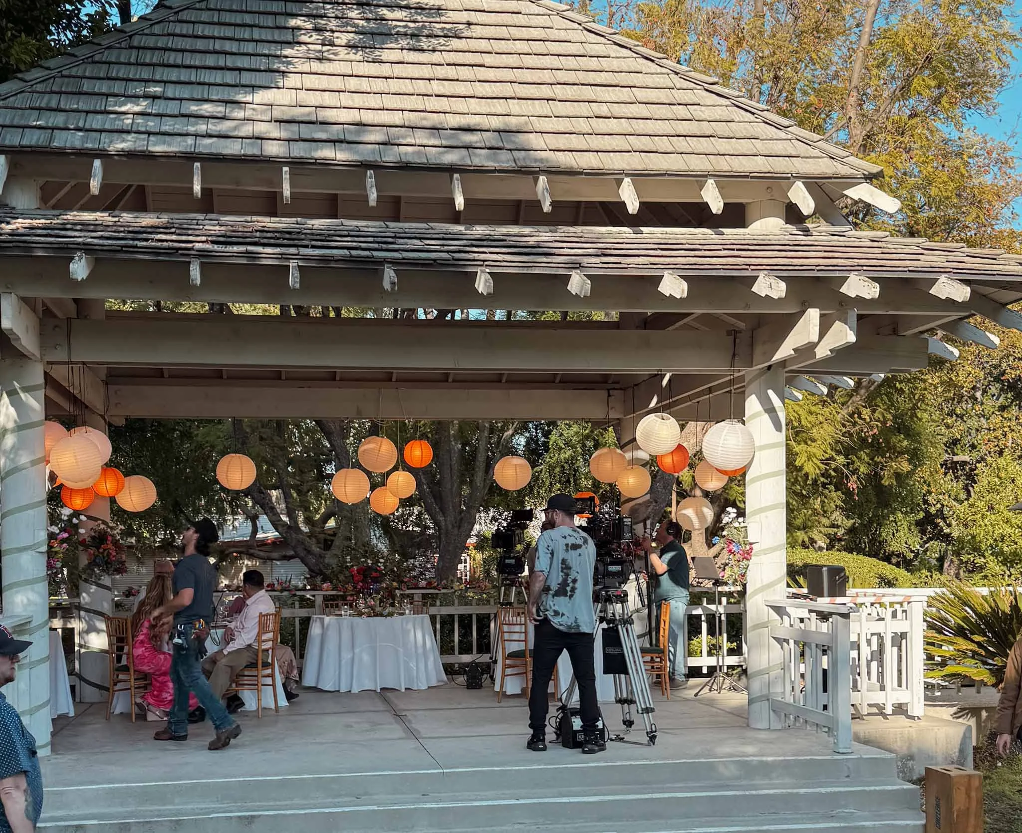 People setting up a video camera in front of a decorated outdoor pavilion with hanging paper lanterns, and tables with white tablecloths.