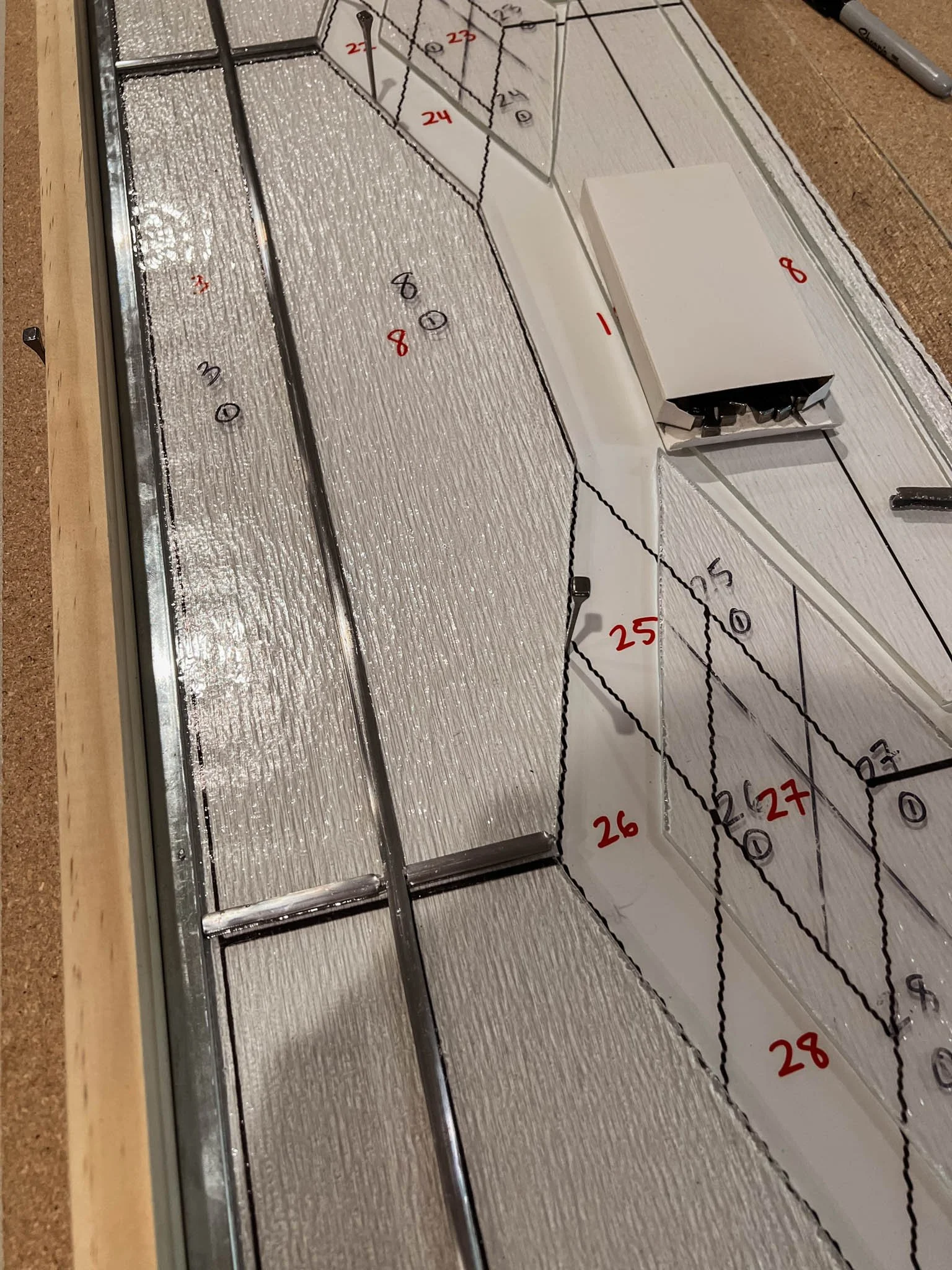 A woodworking assembly with a metal frame, a paper template with numbered outlines, and a white box with a tool inside. The template shows various measurements and marks for cutting or assembly.