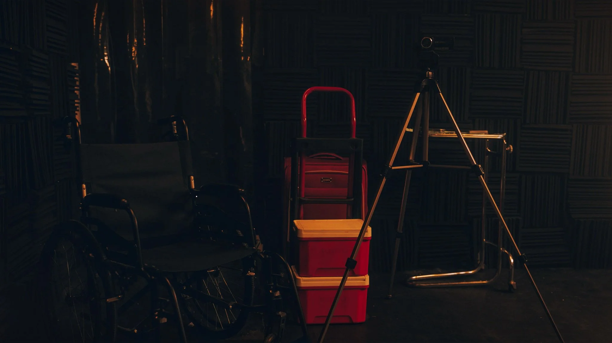 A wheelchair, red and yellow storage bins, a tripod with camera, and a small table against audio foam padded walls in a recording studio or soundproof room.