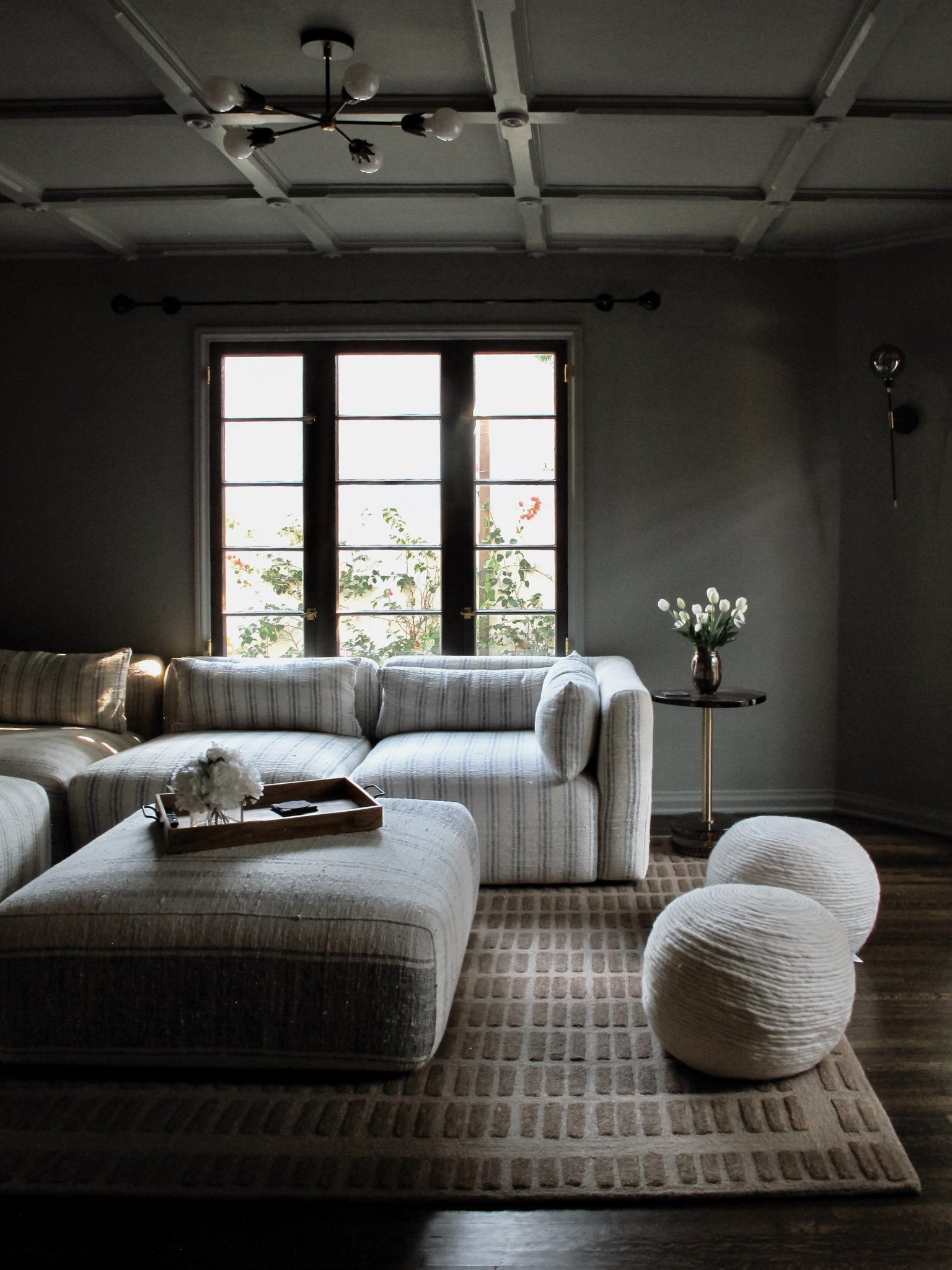 Cozy living room with natural light, featuring a large striped sofa, a matching ottoman with a tray, two white textured poufs, a side table with a vase of white flowers, a patterned rug, and a window with greenery outside.