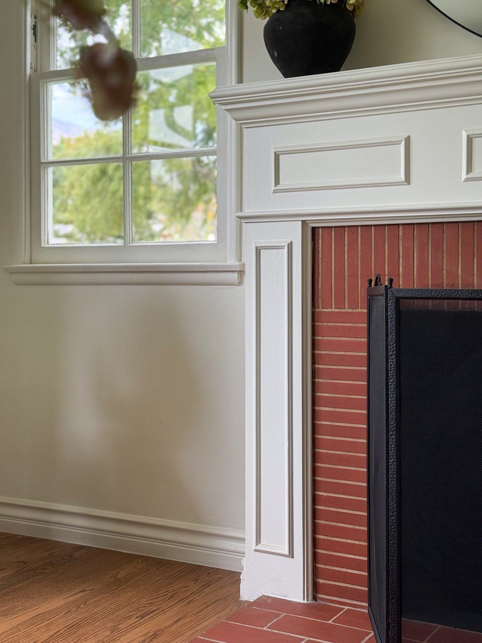Part of a fireplace with a brick surround, white wooden mantle, and black vase on top. A window with multiple panes showing greenery outside. Hardwood floor.