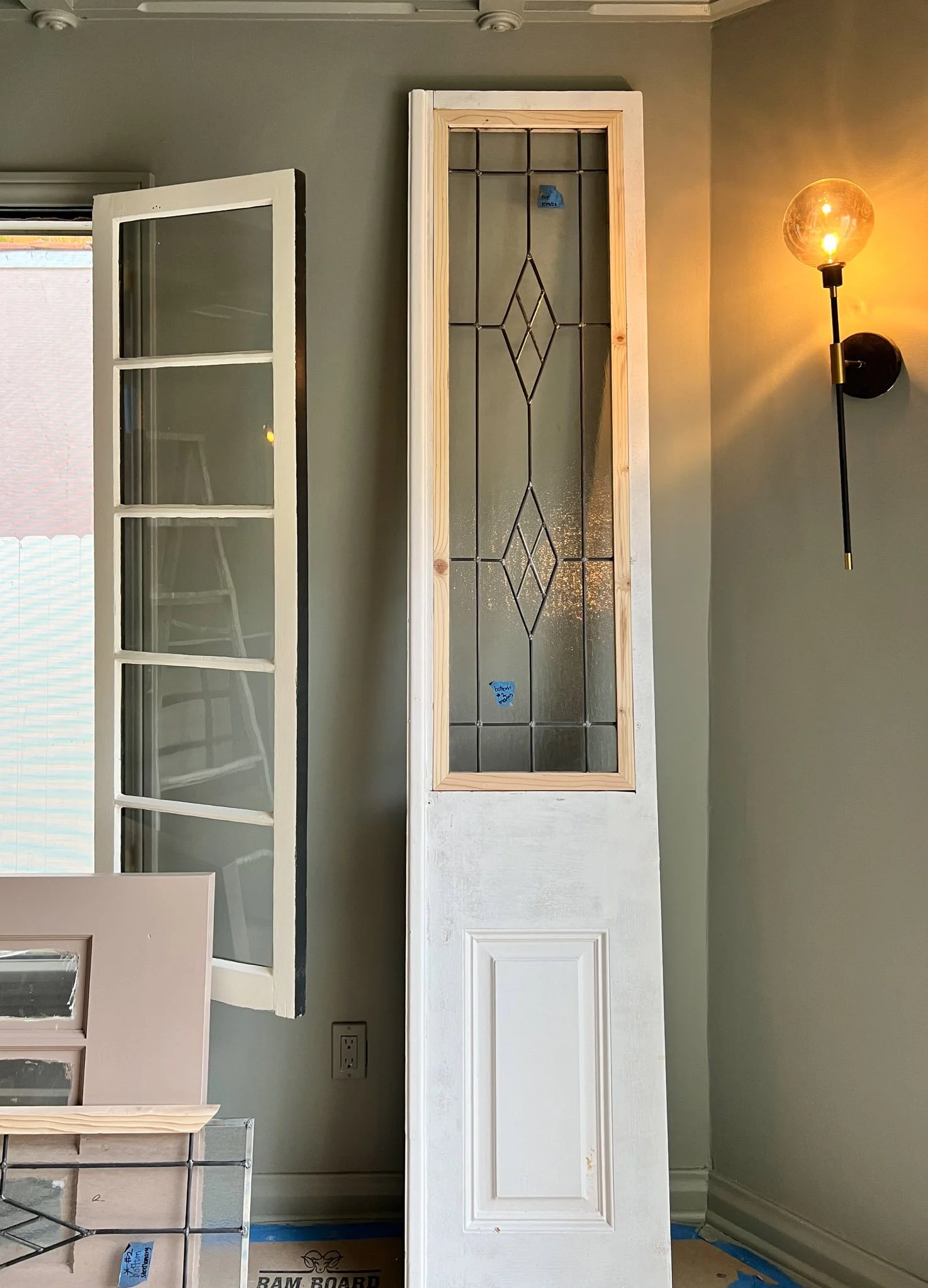 A tall wooden door with a glass panel featuring decorative diamond patterns, leaning against a wall in a room with a gray wall. To the left, a window with a white frame and a glass pane is open, revealing blinds outside. To the right, a wall-mounted 
