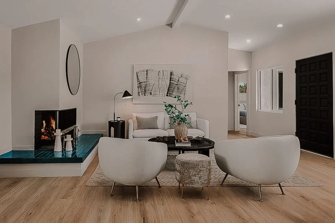 Modern living room with white walls, a fireplace with teal tiles, beige seating, a black coffee table with a vase and plants, and a black front door.