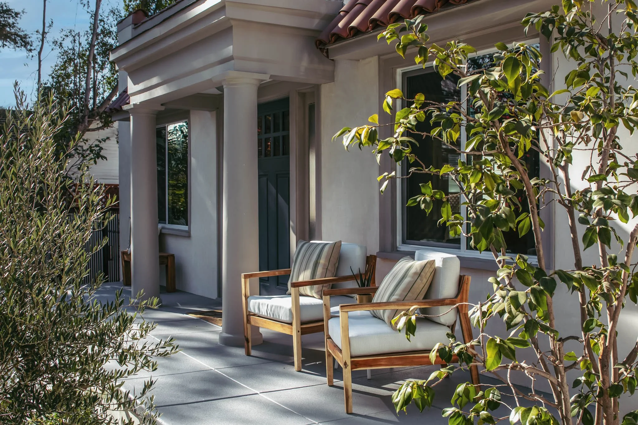 Front porch of a house with two wooden chairs with white cushions and striped pillows, a small side table, and a potted plant, surrounded by greenery.