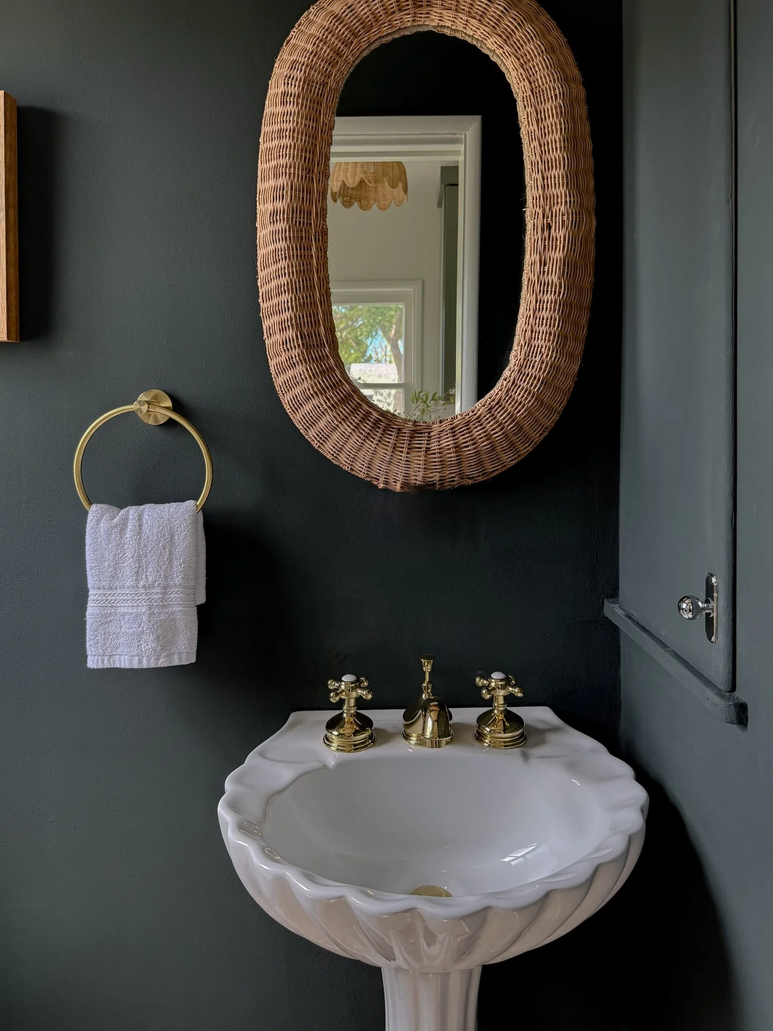 Bathroom with dark green wall, wicker-framed oval mirror, white pedestal sink with gold fixtures, and a white towel on a gold towel ring.