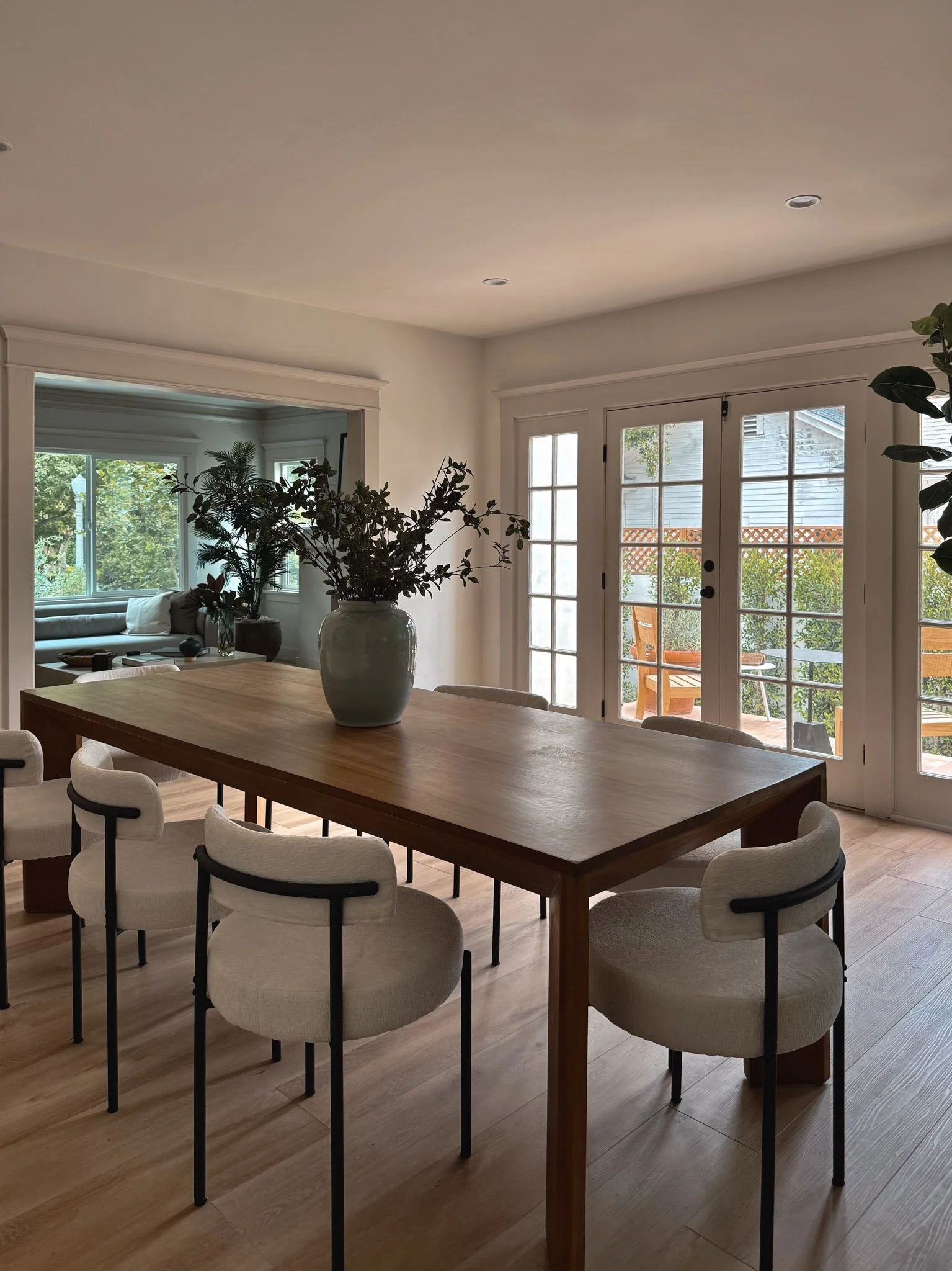 A dining room with a wooden table, light-colored upholstered chairs, a large vase with green foliage on the table, and French doors leading to a patio with outdoor furniture. There's a view into a living room with a sofa and windows.
