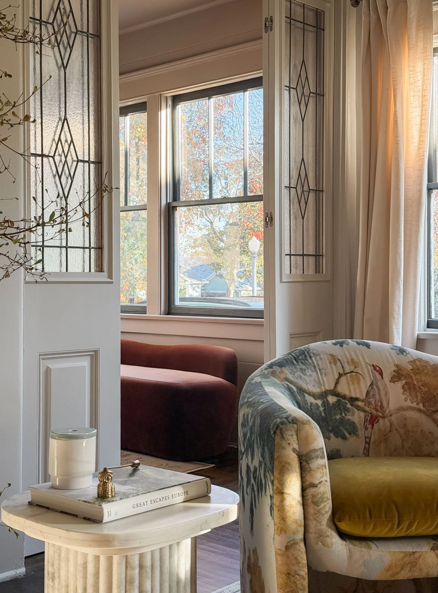A cozy corner of a living room with a floral upholstered armchair and a small round coffee table holding a book titled 'Great Escapes Europe' and a white container, with large windows showing autumn trees outside.
