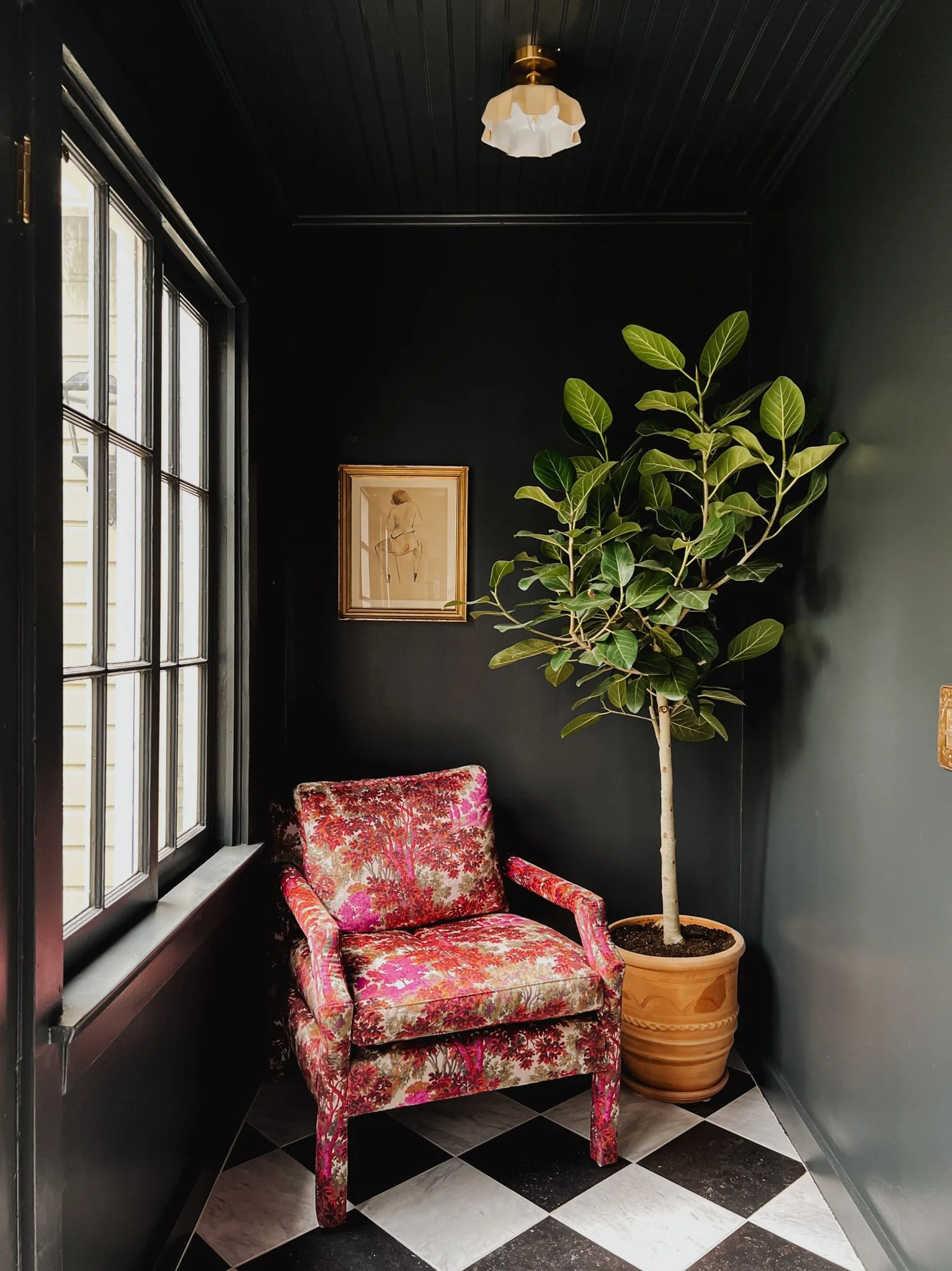 A cozy room corner with a upholstered armchair in bright, floral fabric, a tall potted plant with broad green leaves, a large window with black framing, dark walls, a framed artwork of a woman, and a ceiling light fixture.