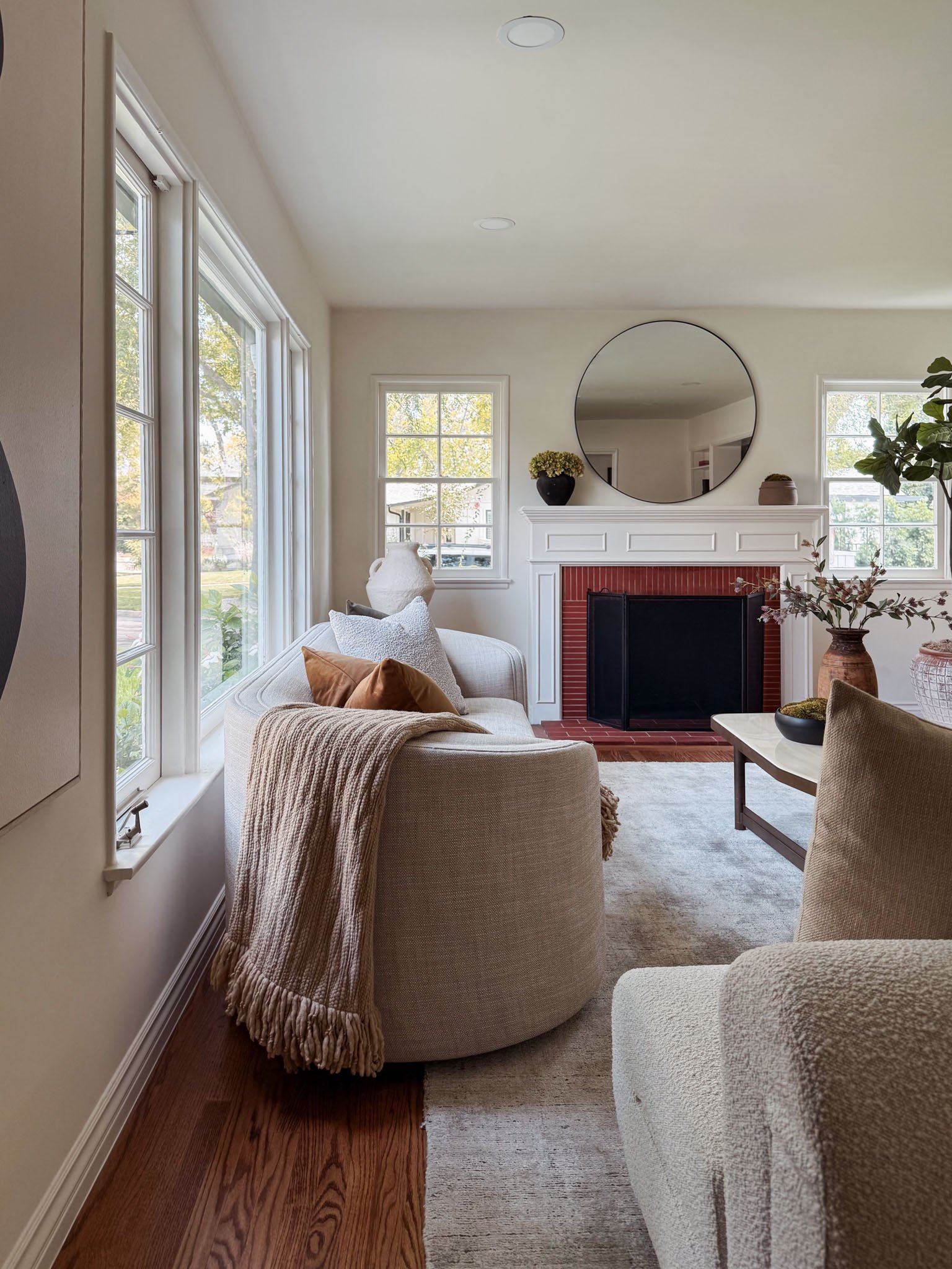 Living room with large windows, white walls, a white couch with throw pillows and a beige blanket, a modern white fireplace with a round mirror above it, decorative vases and plants, and hardwood floors.