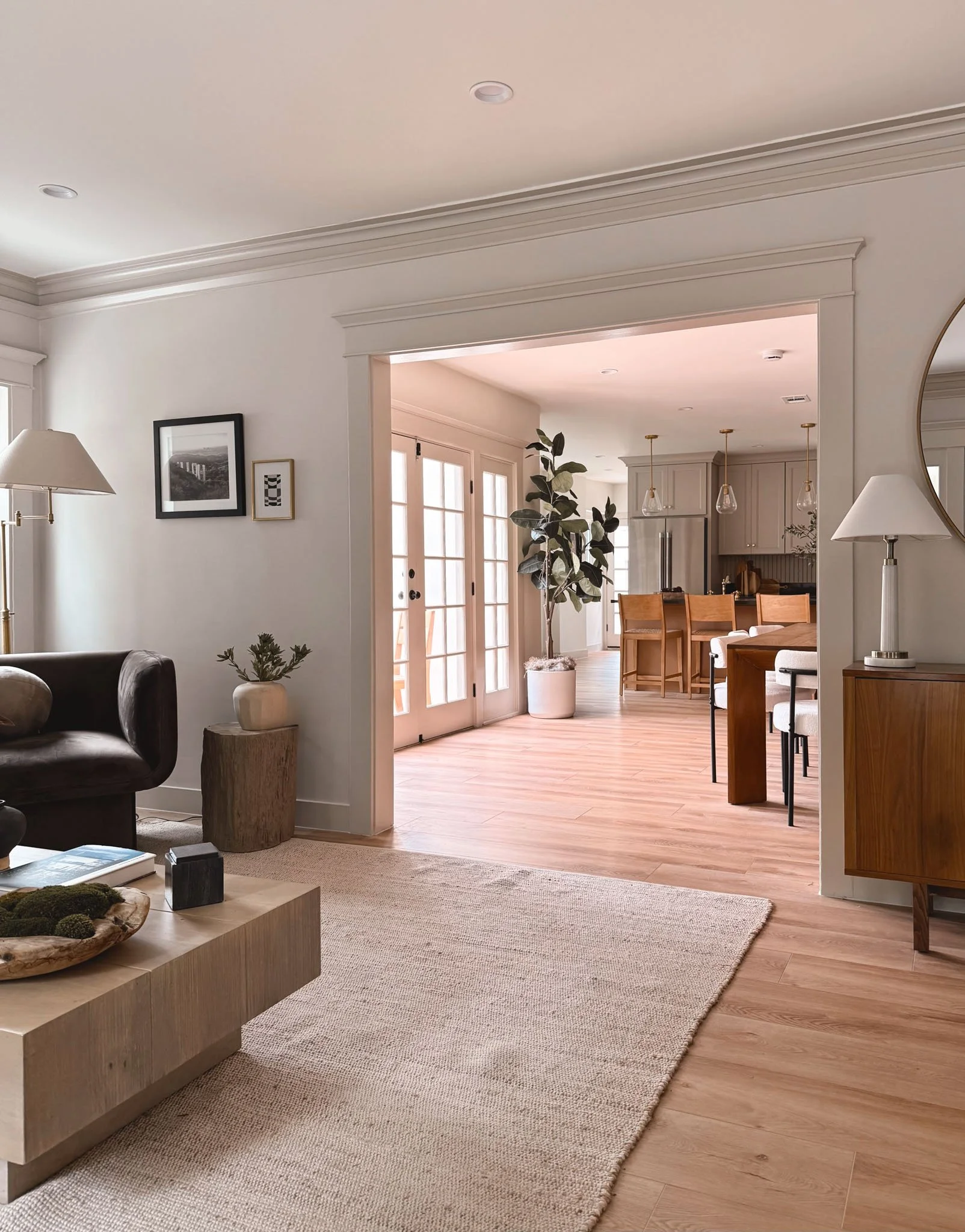 A bright living room with beige walls, a brown leather sofa, a wooden coffee table with decor, a floor lamp, framed pictures, and a doorway leading to a dining area with a large potted plant, wooden chairs, and kitchen in the background.