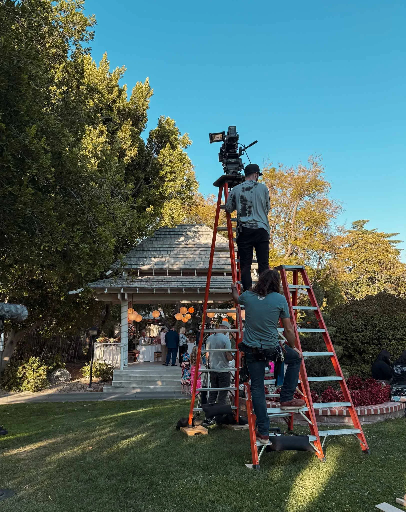 A film crew setting up camera equipment on ladders outside a small pavilion with strings of hanging lanterns, surrounded by trees and bushes during the daytime.