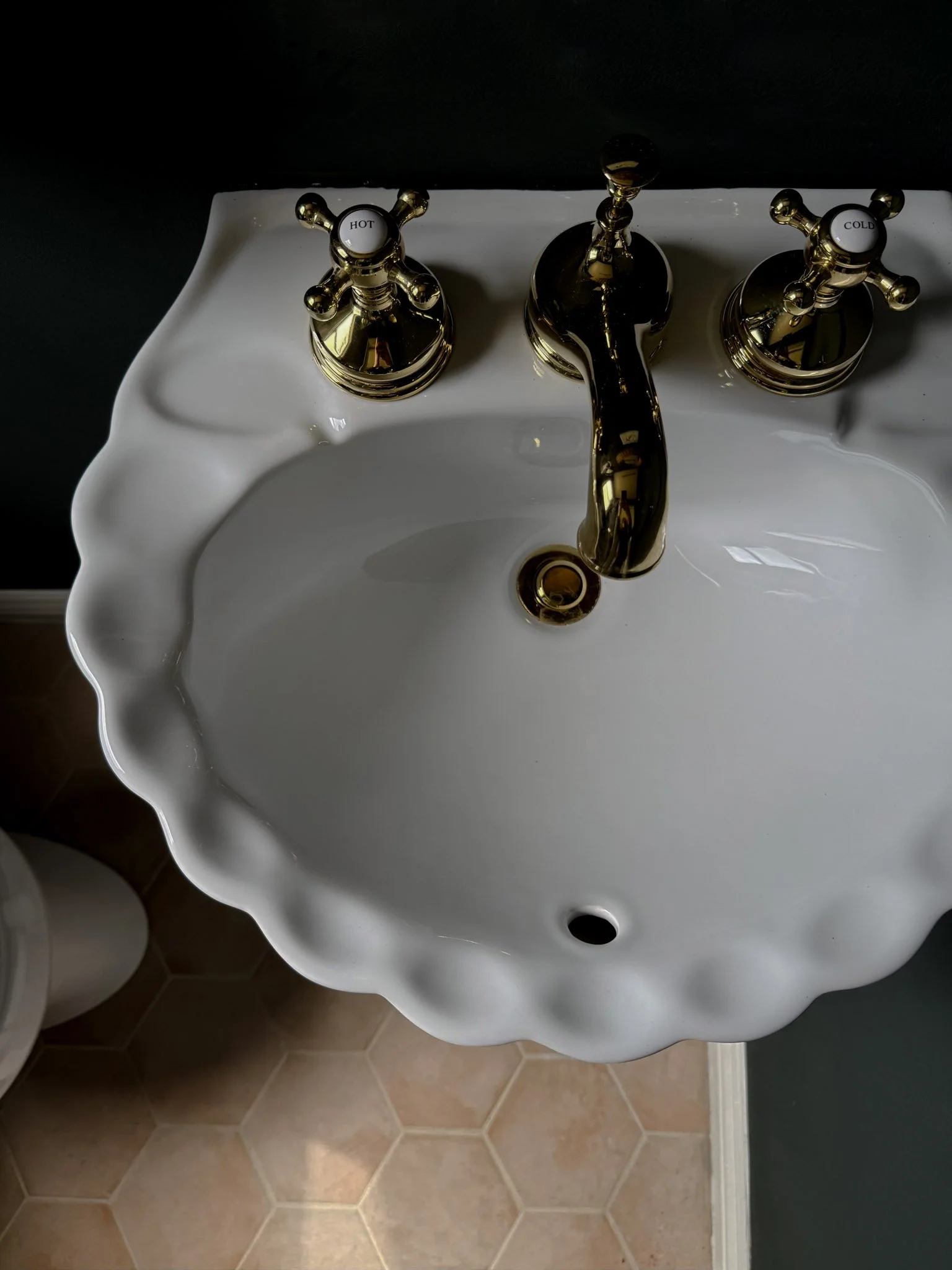 White porcelain bathroom sink with scalloped edge, gold faucet with hot and cold handles, and hexagonal tan tile flooring