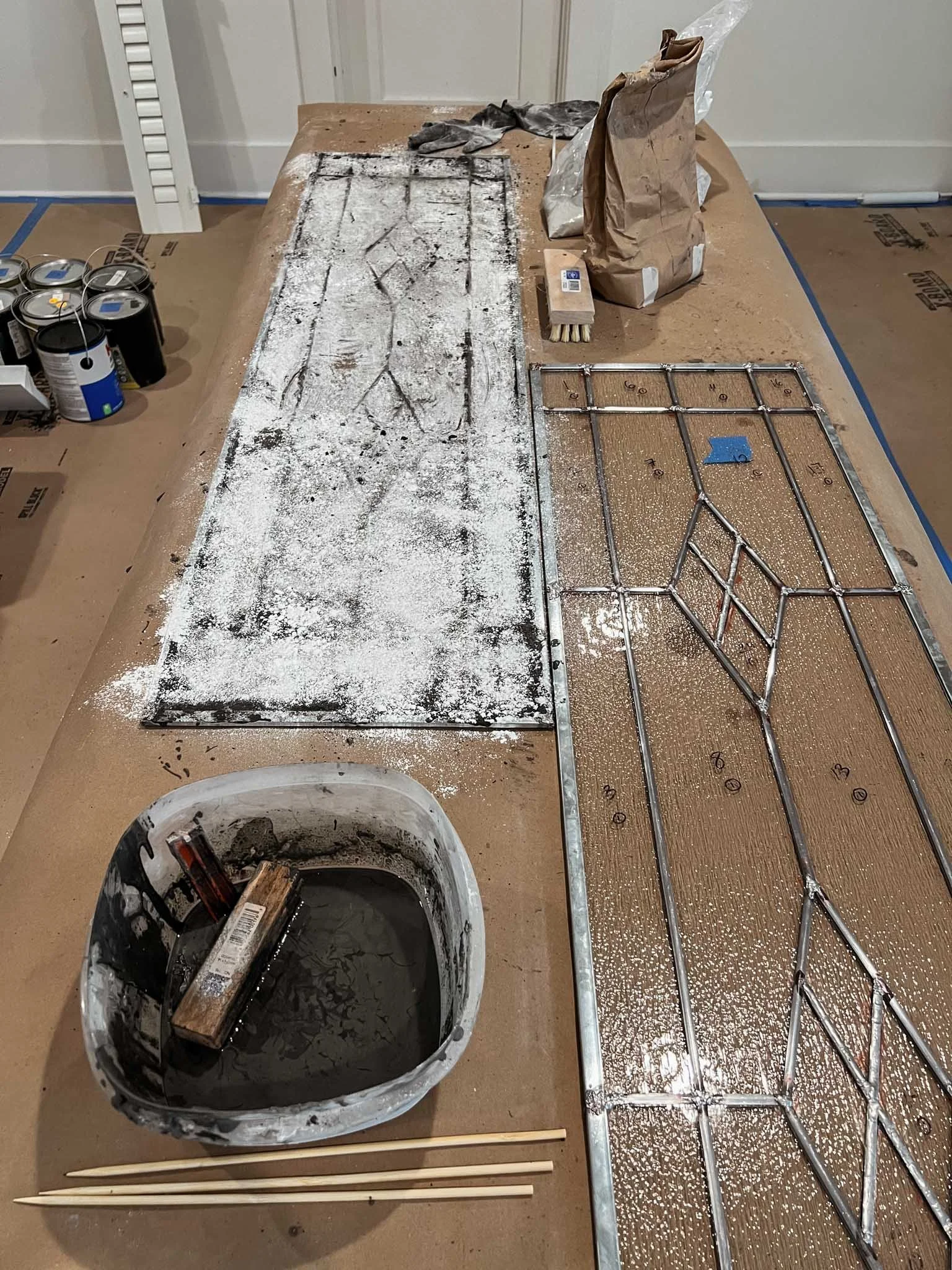 A worktable with disassembled metal framework, a container of water with a brush, and cans of paint or primer, along with bags and tools, in a workspace setting.