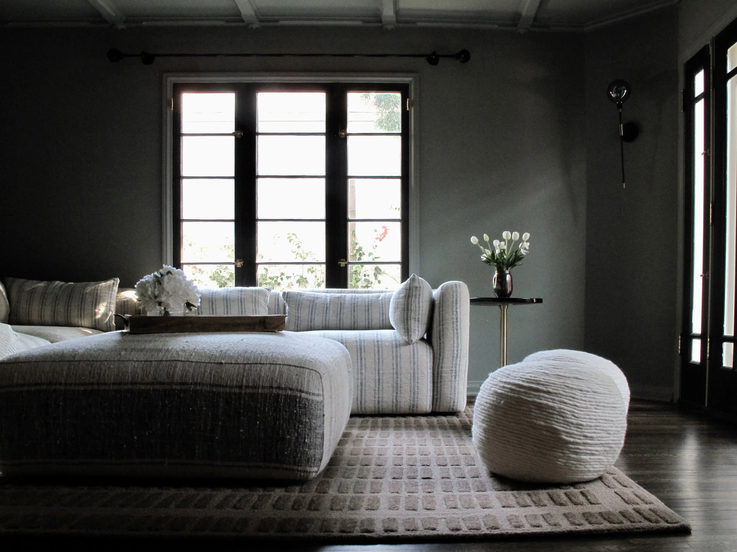 Living room with dark gray walls, large window with black frame, beige striped sofa, white woven pouf, small side table with a vase of white tulips, and wooden floor with beige area rug.