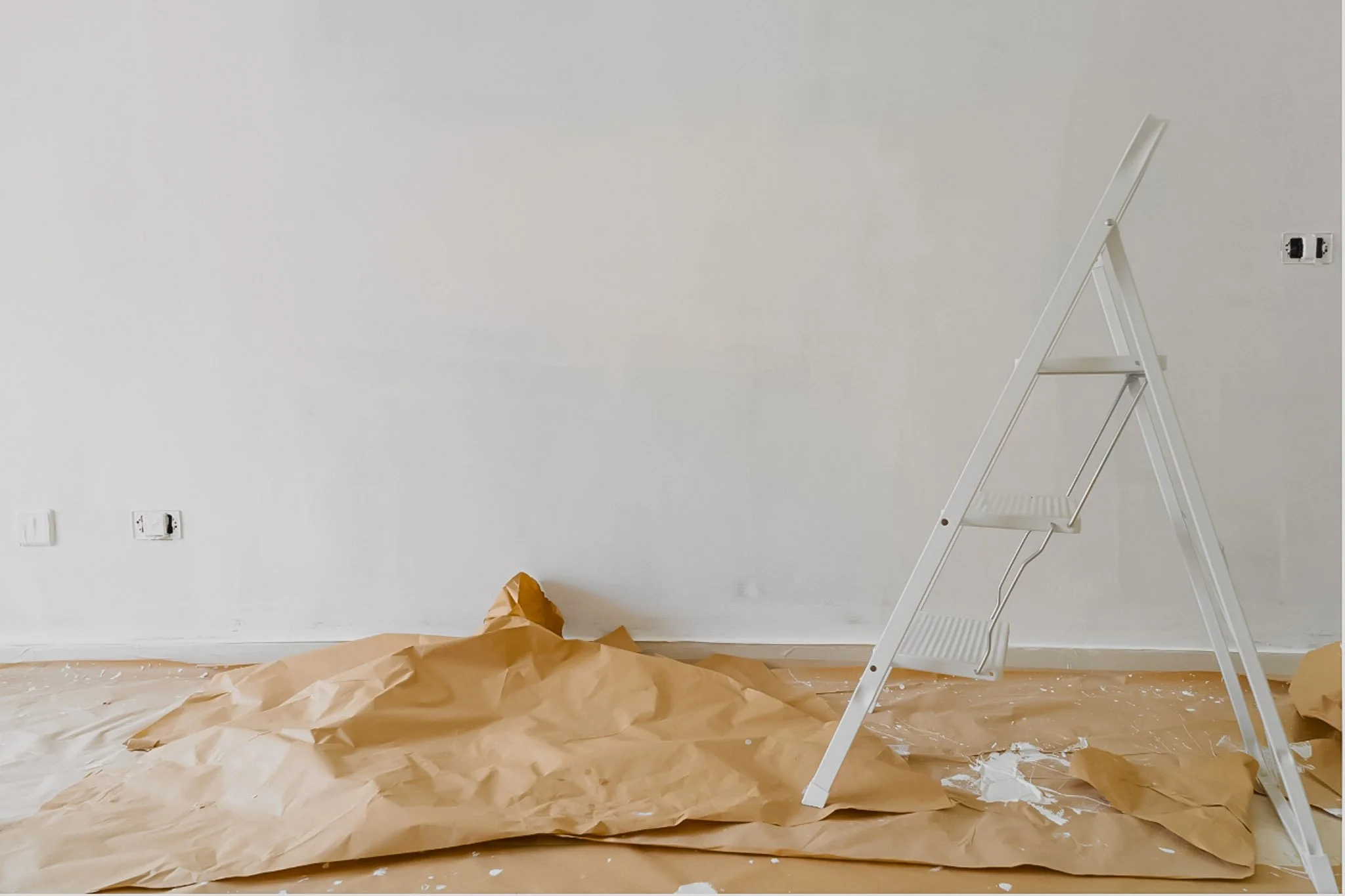 Empty room with a white A-frame ladder, brown paper covering the floor, and paint splatters, indicating painting or renovation work.