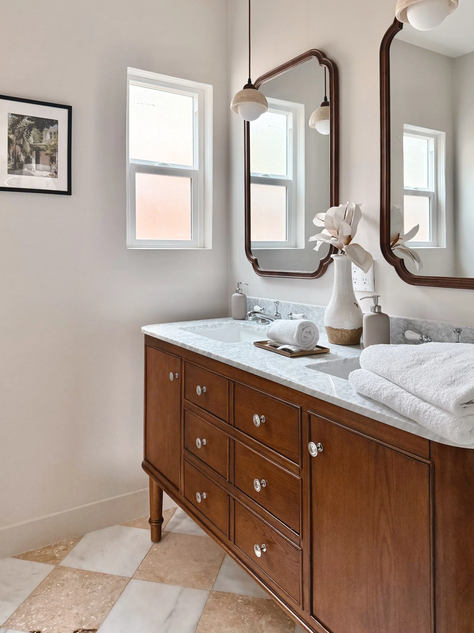 Interior of a bathroom with a wooden vanity with a marble countertop, two mirrors, a vase with flowers, folded towels, and two windows with frosted glass panes.
