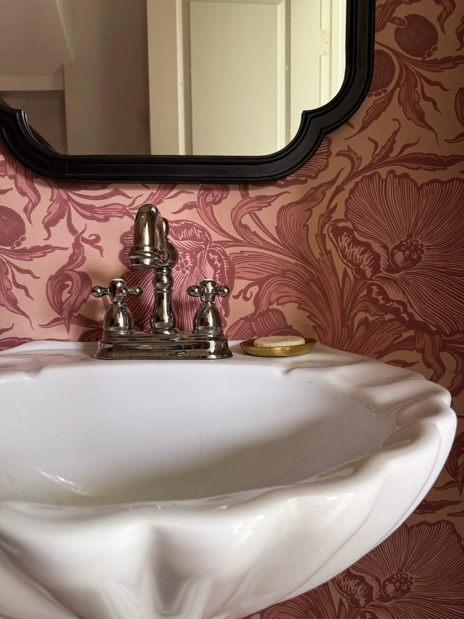 A white porcelain vintage bathroom sink with a chrome faucet, a soap dish with soap, a black-framed mirror, and wallpaper with pink floral pattern.
